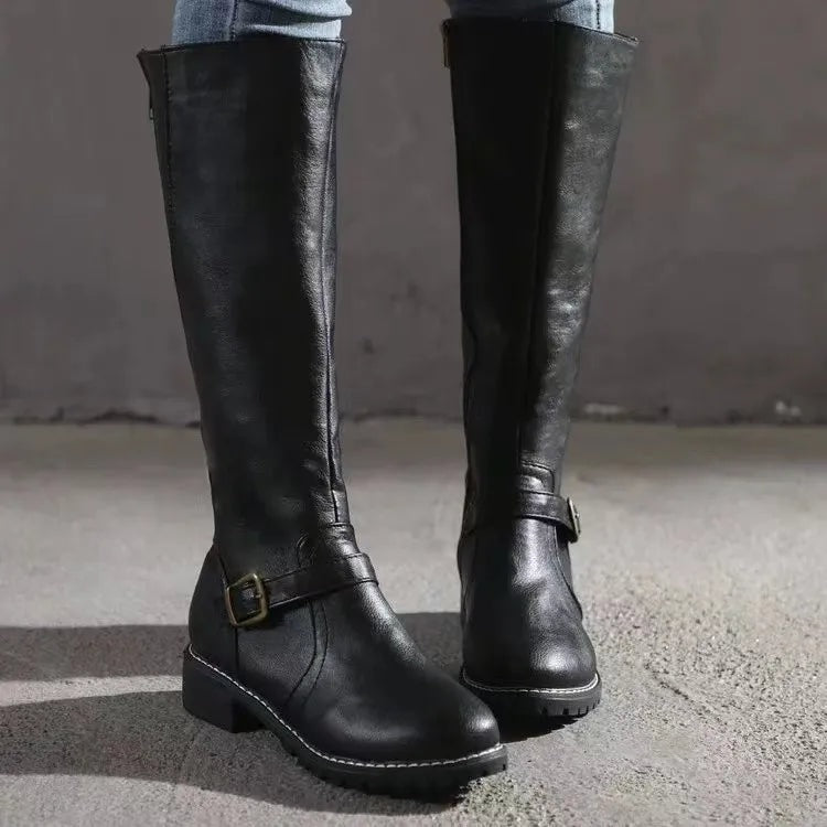 Black knee-high boots standing side by side on pavement, showing the tall shafts, ankle buckles, low heels, and textured soles.