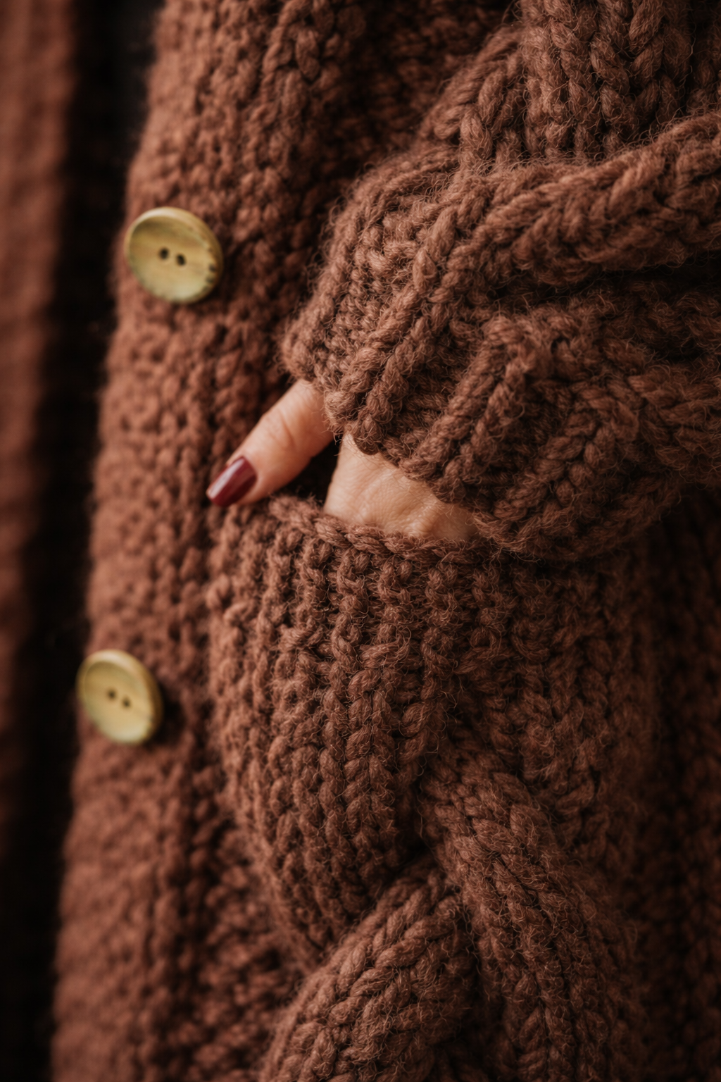 Close-up detail of the brown cardigan coat showing its textured cable knit, button front, and hand resting inside a side pocket.