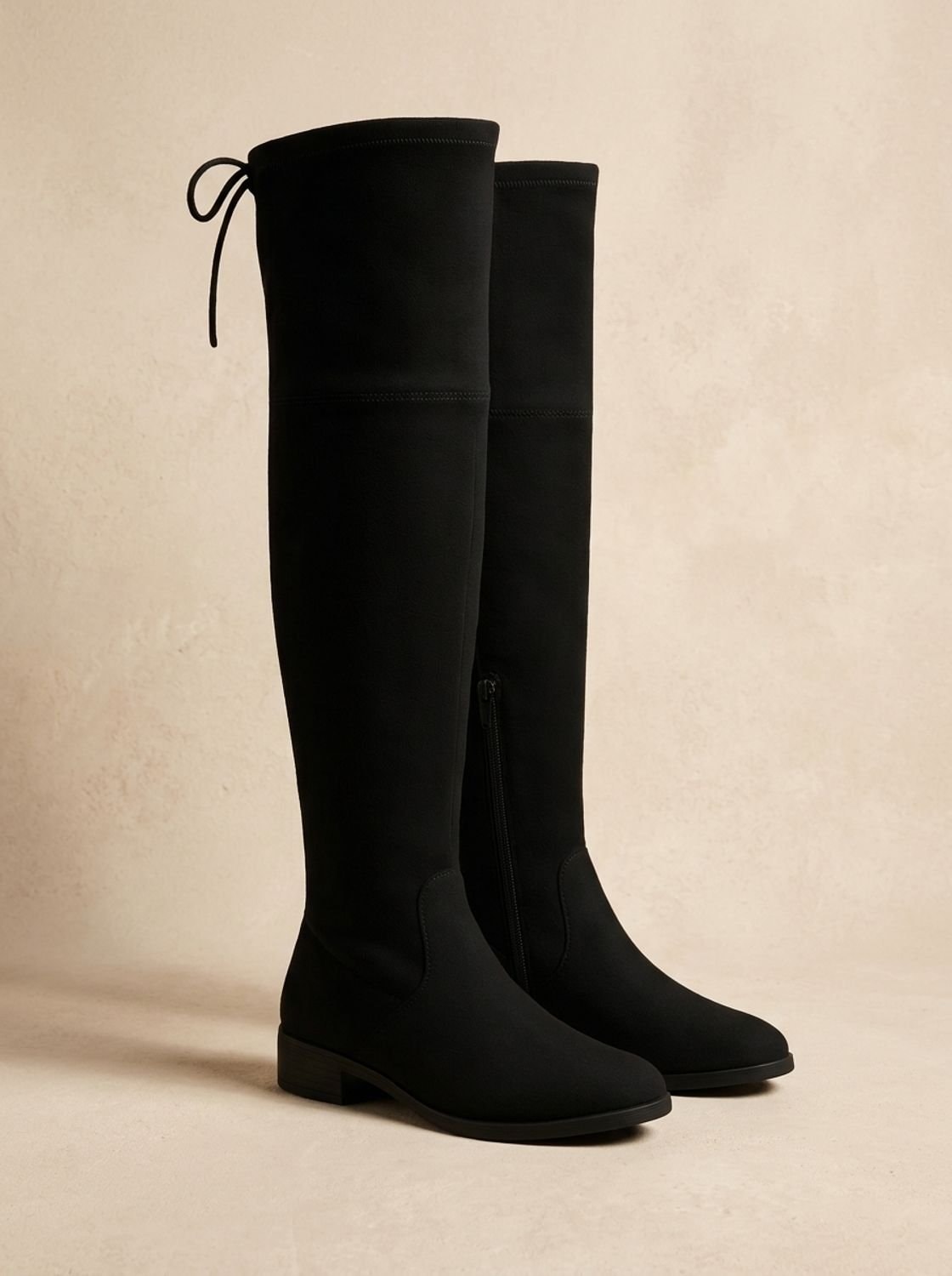 Black over-the-knee flat boots shown in profile, featuring sleek tall shafts, back ties, rounded toes, and low heels.