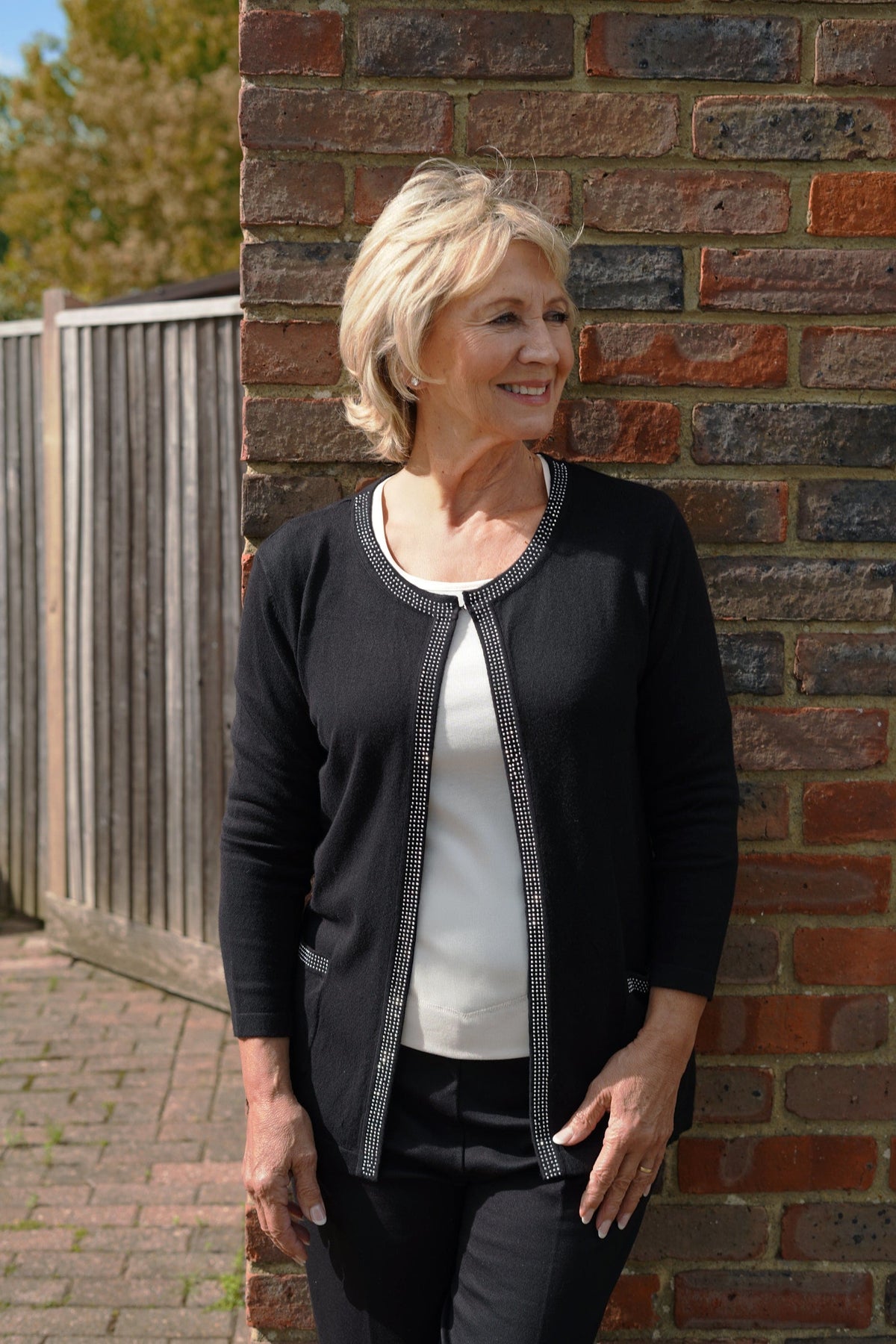 Woman wearing a longline open-front cardigan with studded trim along the neckline and front edges, over a plain top and trousers._0