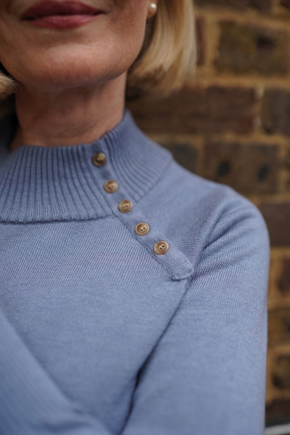 Woman wearing a long sleeve knitted jumper with a funnel collar, buttons along one side of the neck and buttoned ribbed cuffs._12