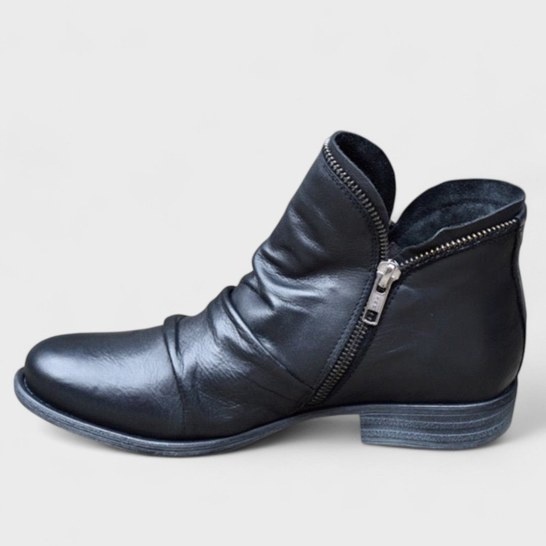 Black ankle boot in side view highlighting its curved side zip, softly gathered upper, rounded toe, and stacked low heel.