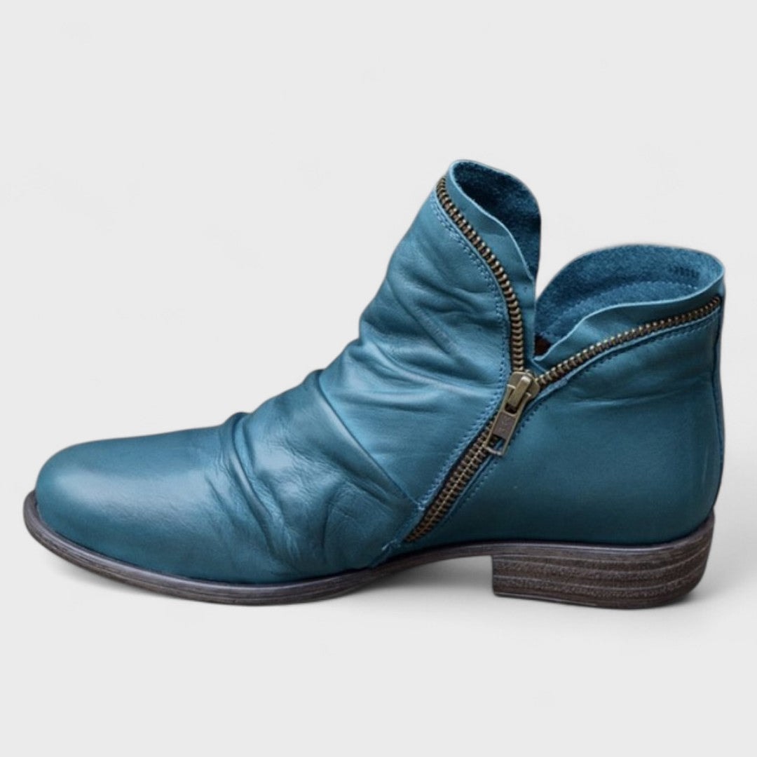 Teal ankle boot displayed from the side with a prominent curved zipper, textured gathered front, rounded toe, and low brown heel.