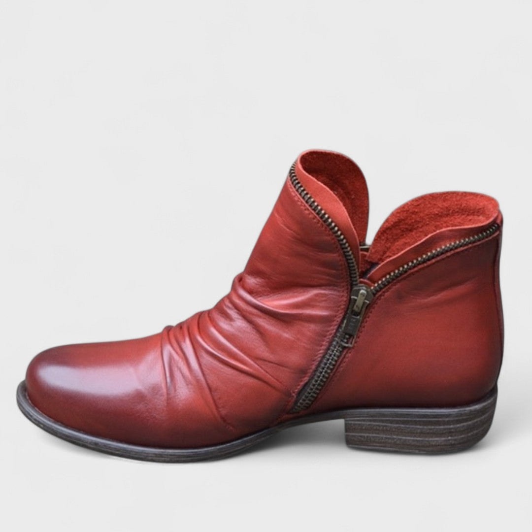 Red ankle boot shown in profile, featuring a curved zip detail, gathered folds across the front, rounded toe, and low block heel.