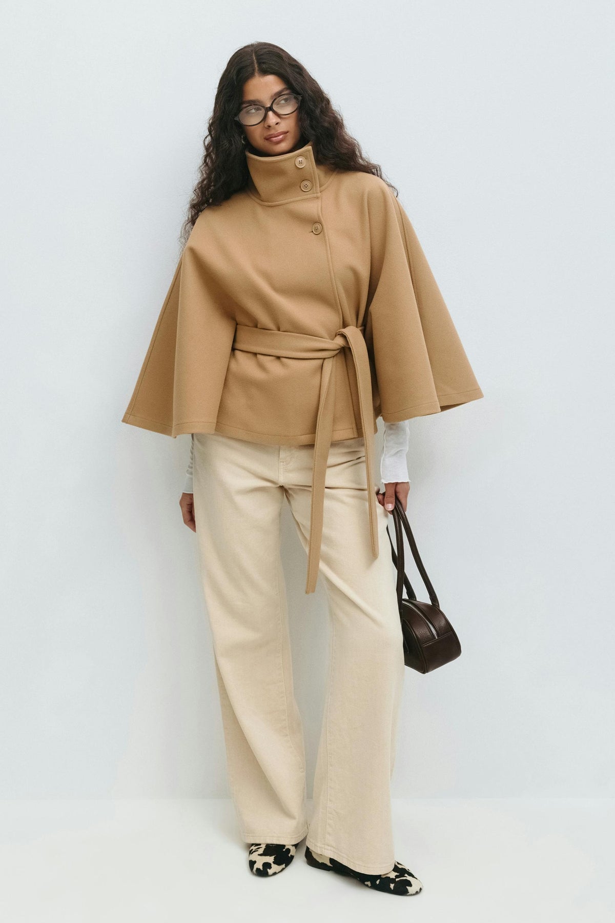 Full-length styling of the camel cape jacket with light trousers, a handbag, and the adjustable belt tied at the side.