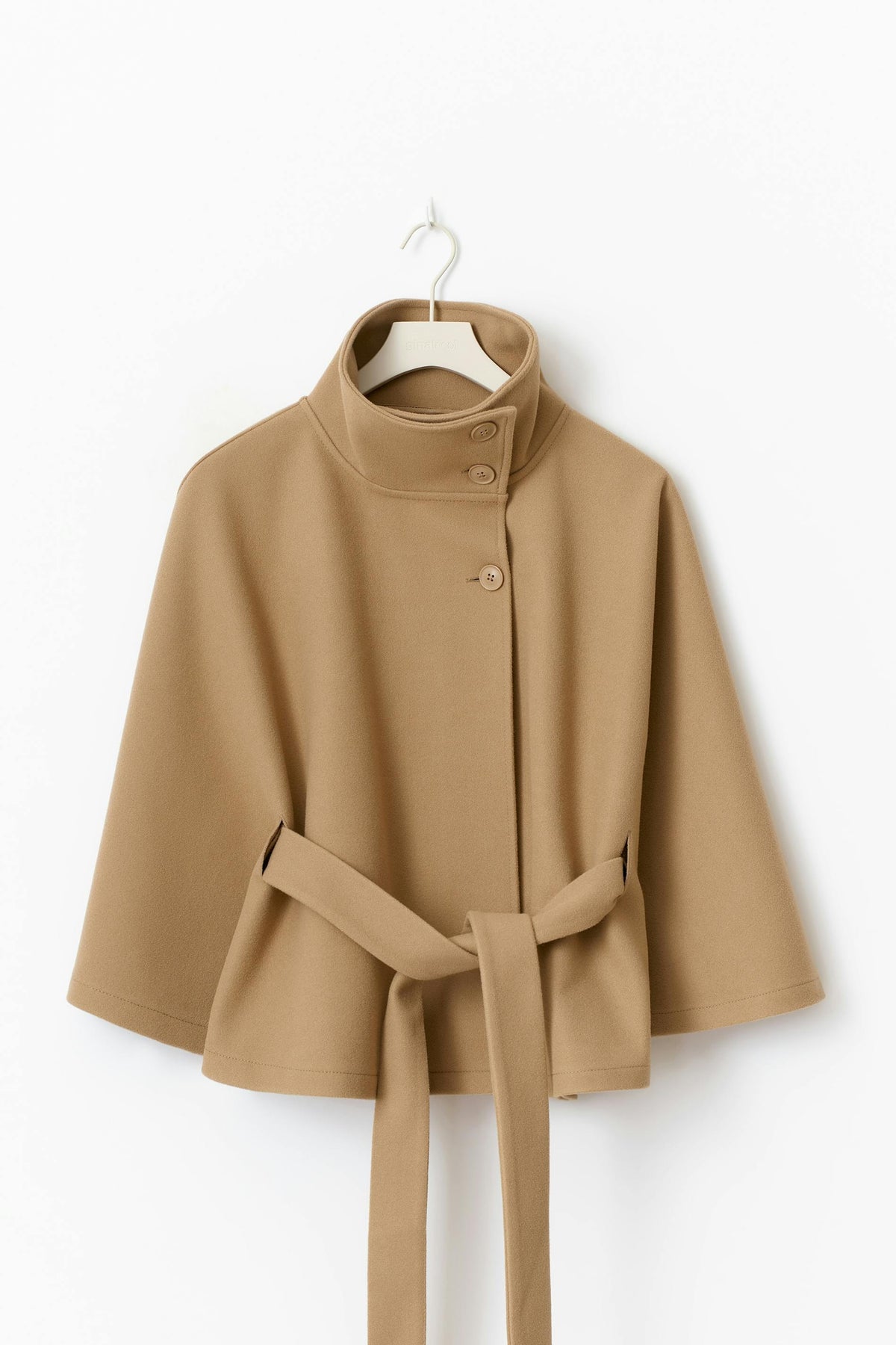 Camel cape jacket displayed on a hanger, showing the stand collar, front buttons, flared sleeves, and belt threaded at the waist.