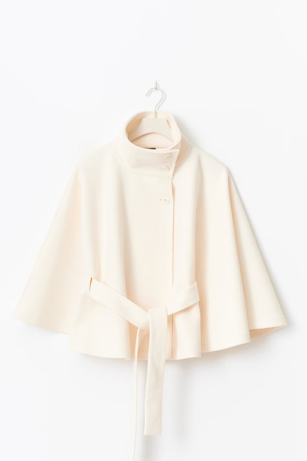 Ivory cape jacket displayed on a hanger with its stand collar, front closure, flared cape sleeves, and loose waist belt visible.