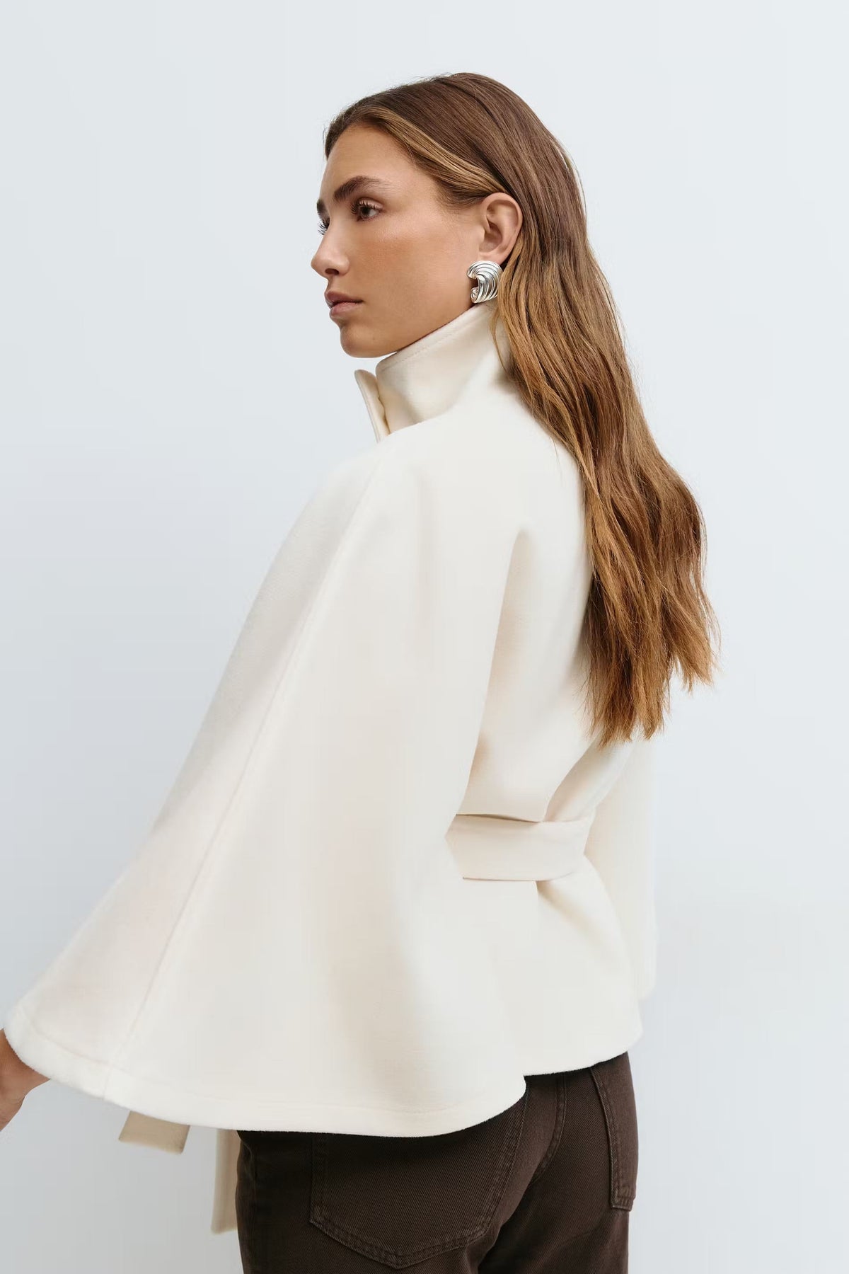 Side-back view of the ivory cape jacket, highlighting the sweeping cape sleeve and waist belt tied behind the model.
