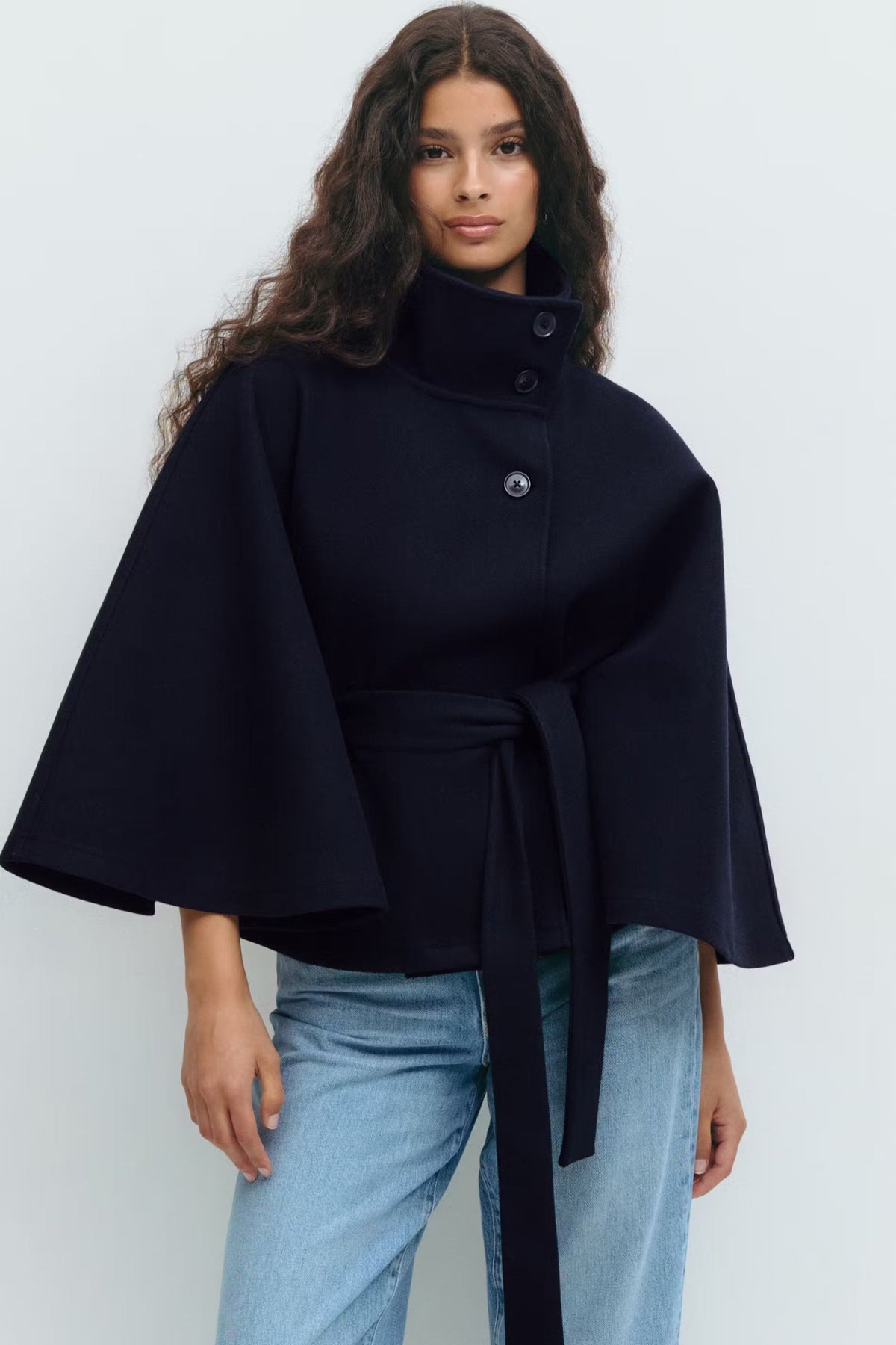 Navy belted cape jacket worn with jeans, featuring a high stand collar, buttoned front, and dramatic cape sleeves.