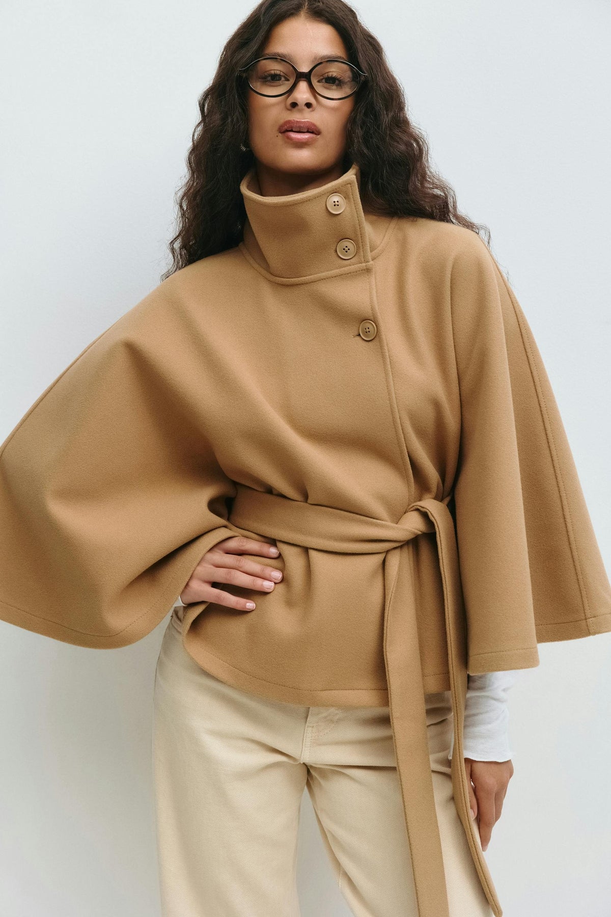 Camel belted cape jacket worn with light trousers, highlighting the structured collar, front buttons, and wide sleeves.