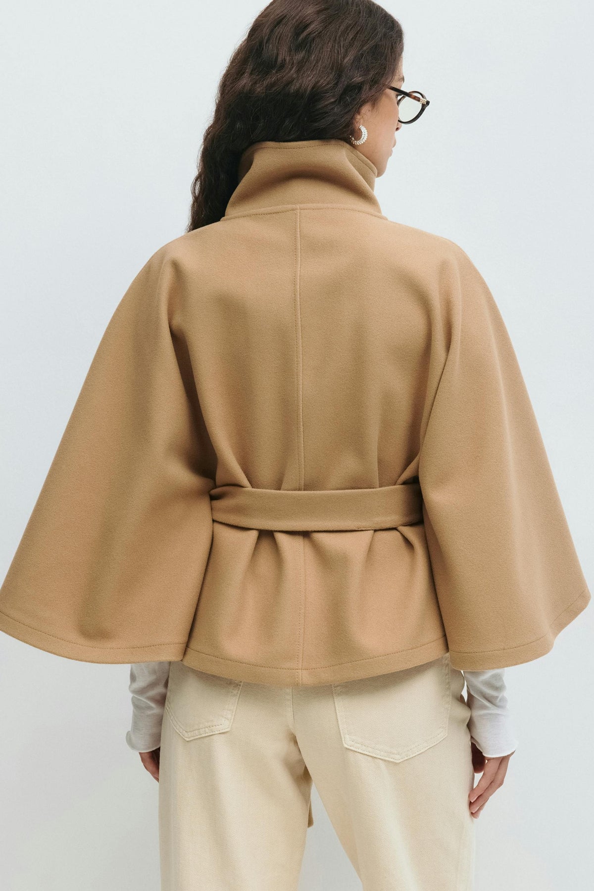 Back view of the camel cape jacket, showing the center seam, broad sleeves, short hem, and belt wrapped across the waist.