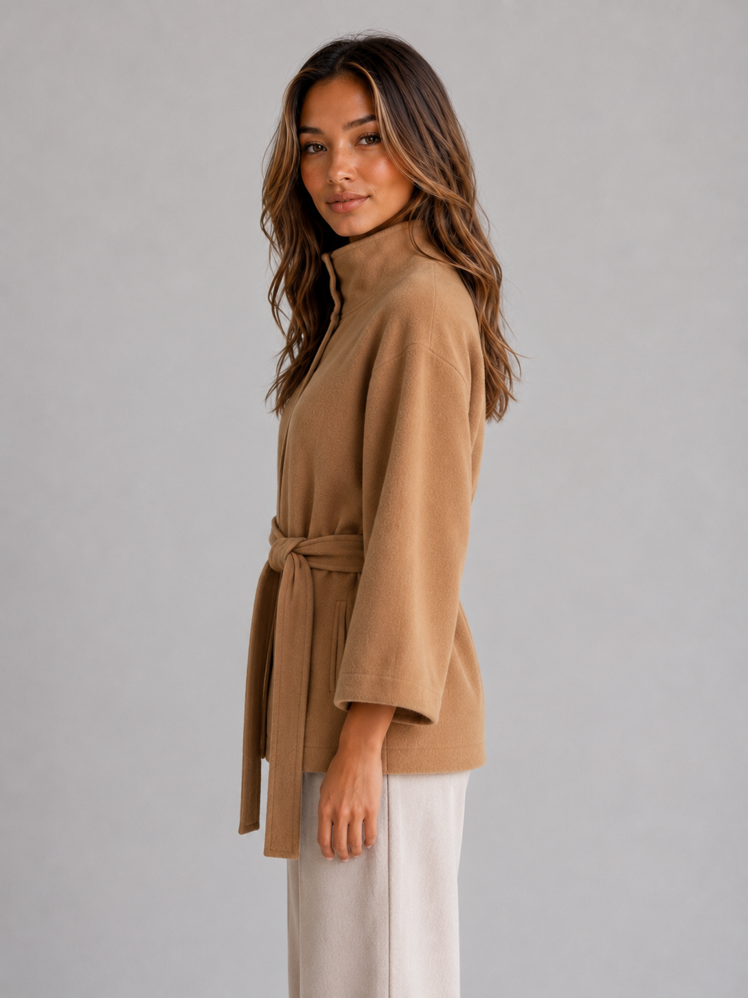 Side profile of the camel belted cape jacket, highlighting its raised collar, flowing sleeve, and waist tie with long ends.