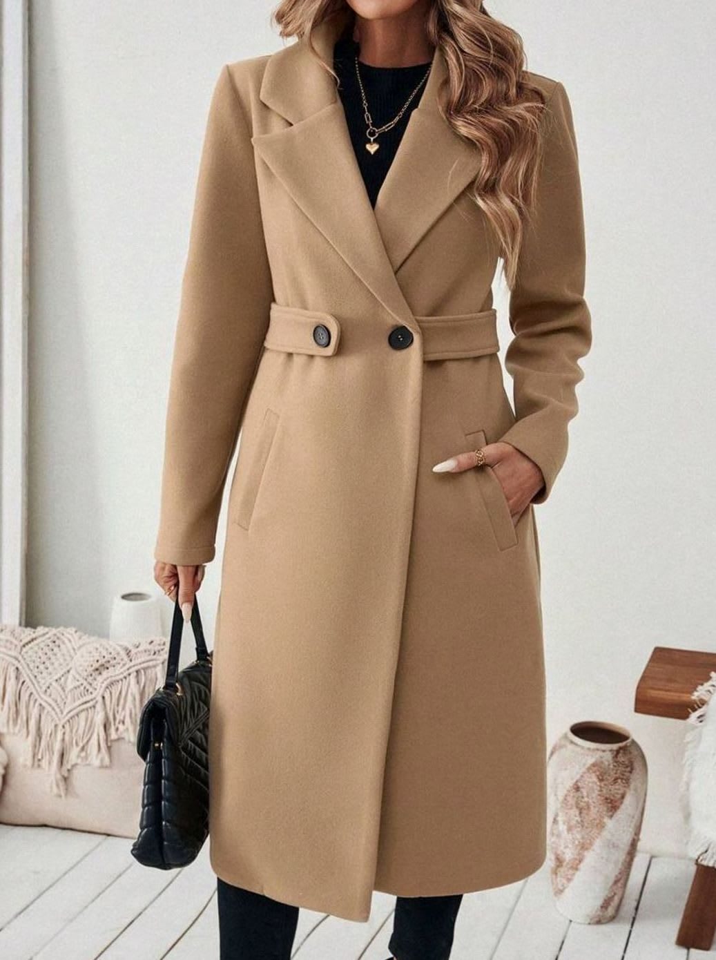 Woman wearing a long notch lapel coat with a buttoned waist tab, welt pockets and long sleeves, holding a quilted handbag._0
