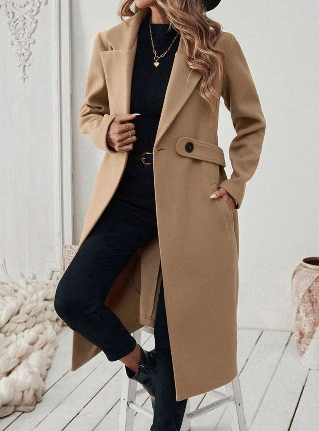 Woman wearing a long notch lapel coat with a buttoned waist tab, welt pockets and long sleeves, holding a quilted handbag._1