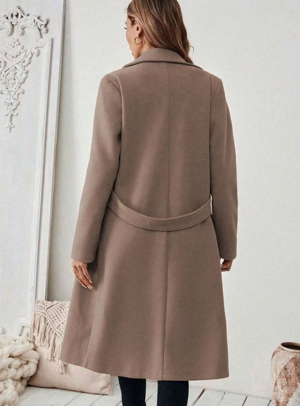 Woman wearing a long notch lapel coat with a buttoned waist tab, welt pockets and long sleeves, holding a quilted handbag._10