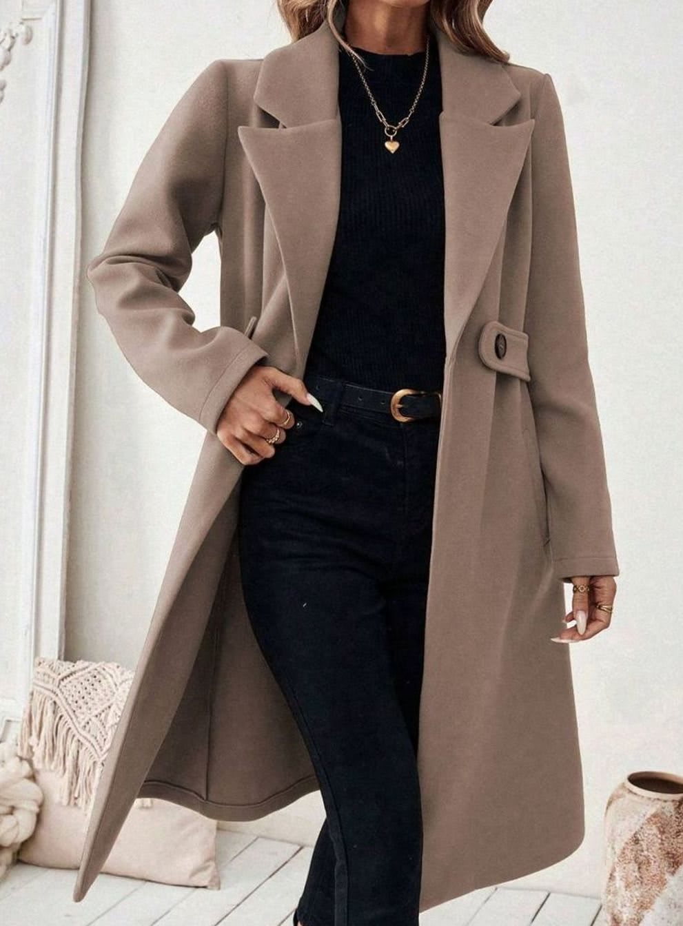Woman wearing a long notch lapel coat with a buttoned waist tab, welt pockets and long sleeves, holding a quilted handbag._11