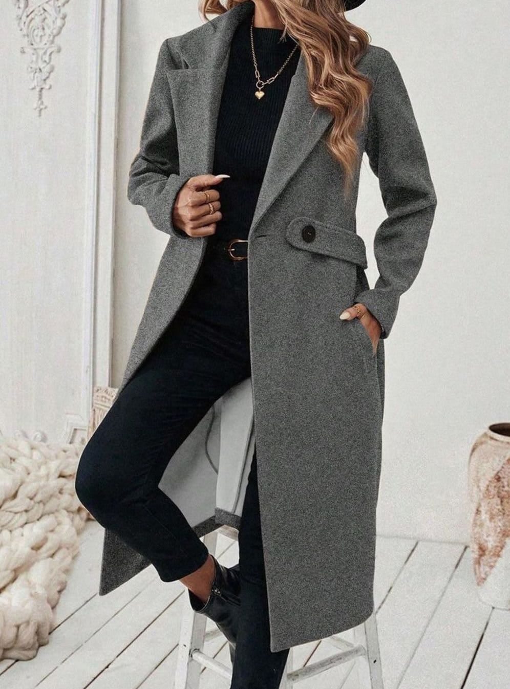 Woman wearing a long notch lapel coat with a buttoned waist tab, welt pockets and long sleeves, holding a quilted handbag._13