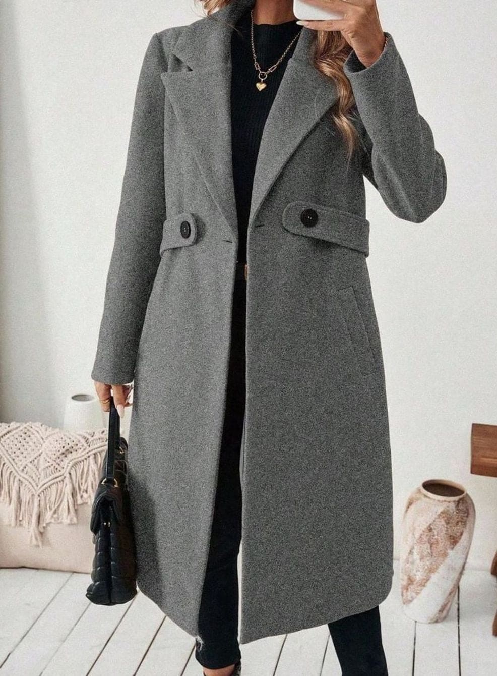 Woman wearing a long notch lapel coat with a buttoned waist tab, welt pockets and long sleeves, holding a quilted handbag._15