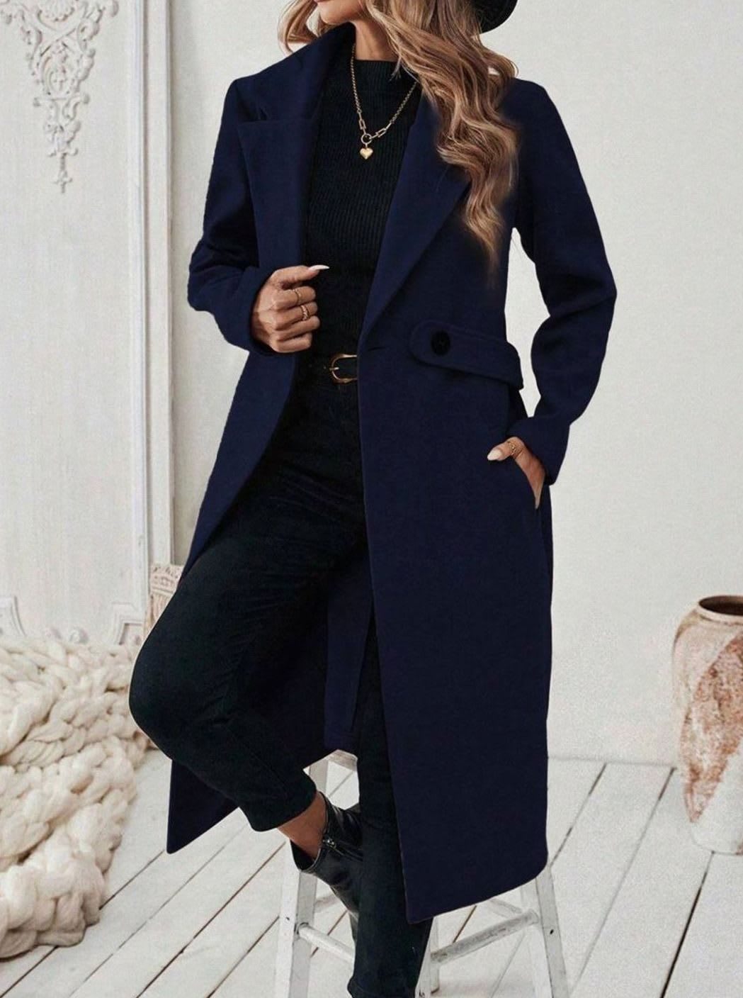 Woman wearing a long notch lapel coat with a buttoned waist tab, welt pockets and long sleeves, holding a quilted handbag._17