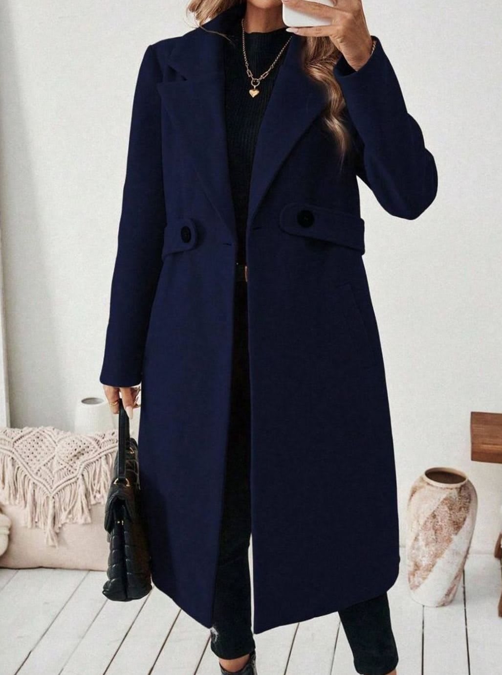 Woman wearing a long notch lapel coat with a buttoned waist tab, welt pockets and long sleeves, holding a quilted handbag._19