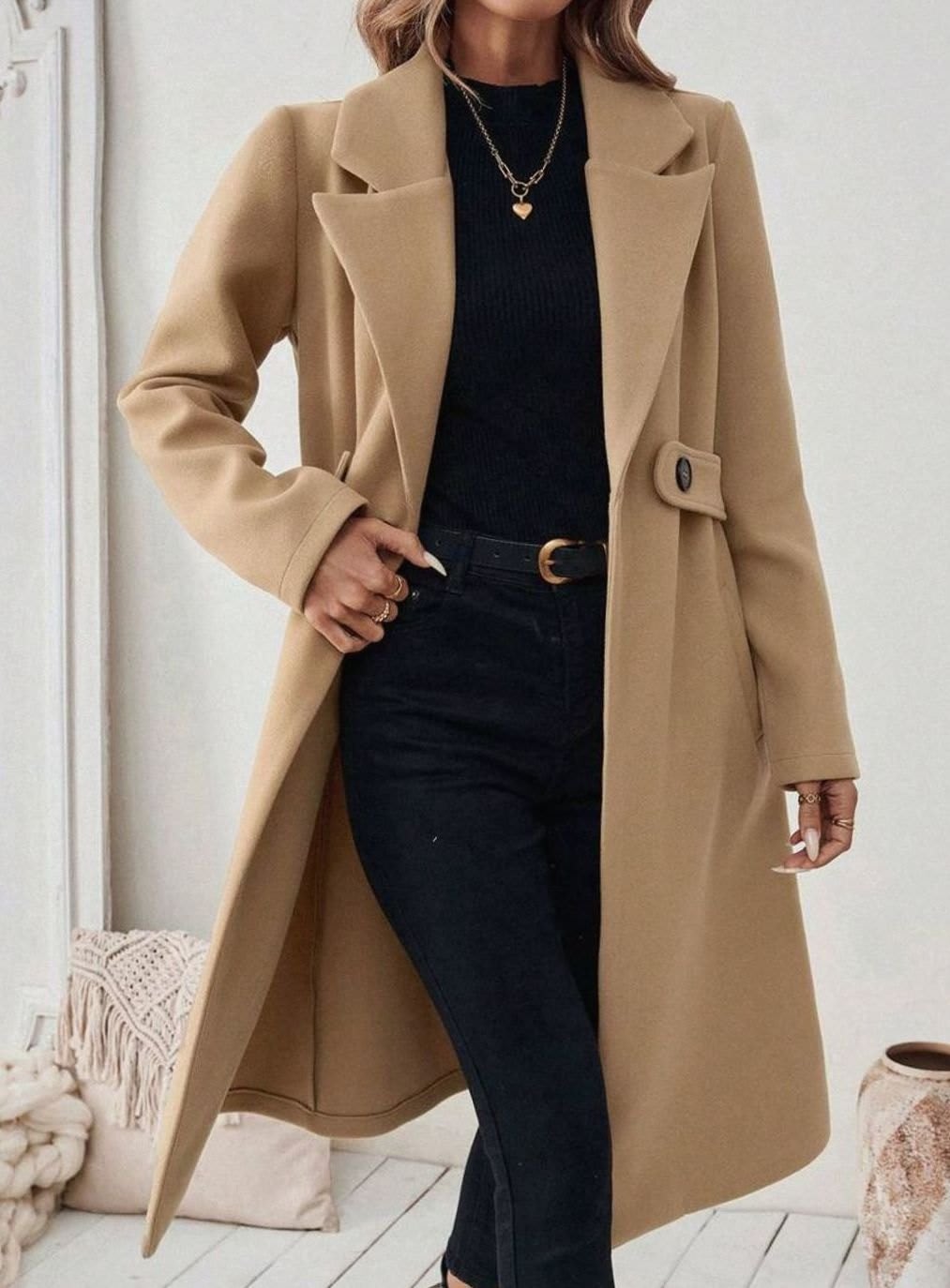 Woman wearing a long notch lapel coat with a buttoned waist tab, welt pockets and long sleeves, holding a quilted handbag._3
