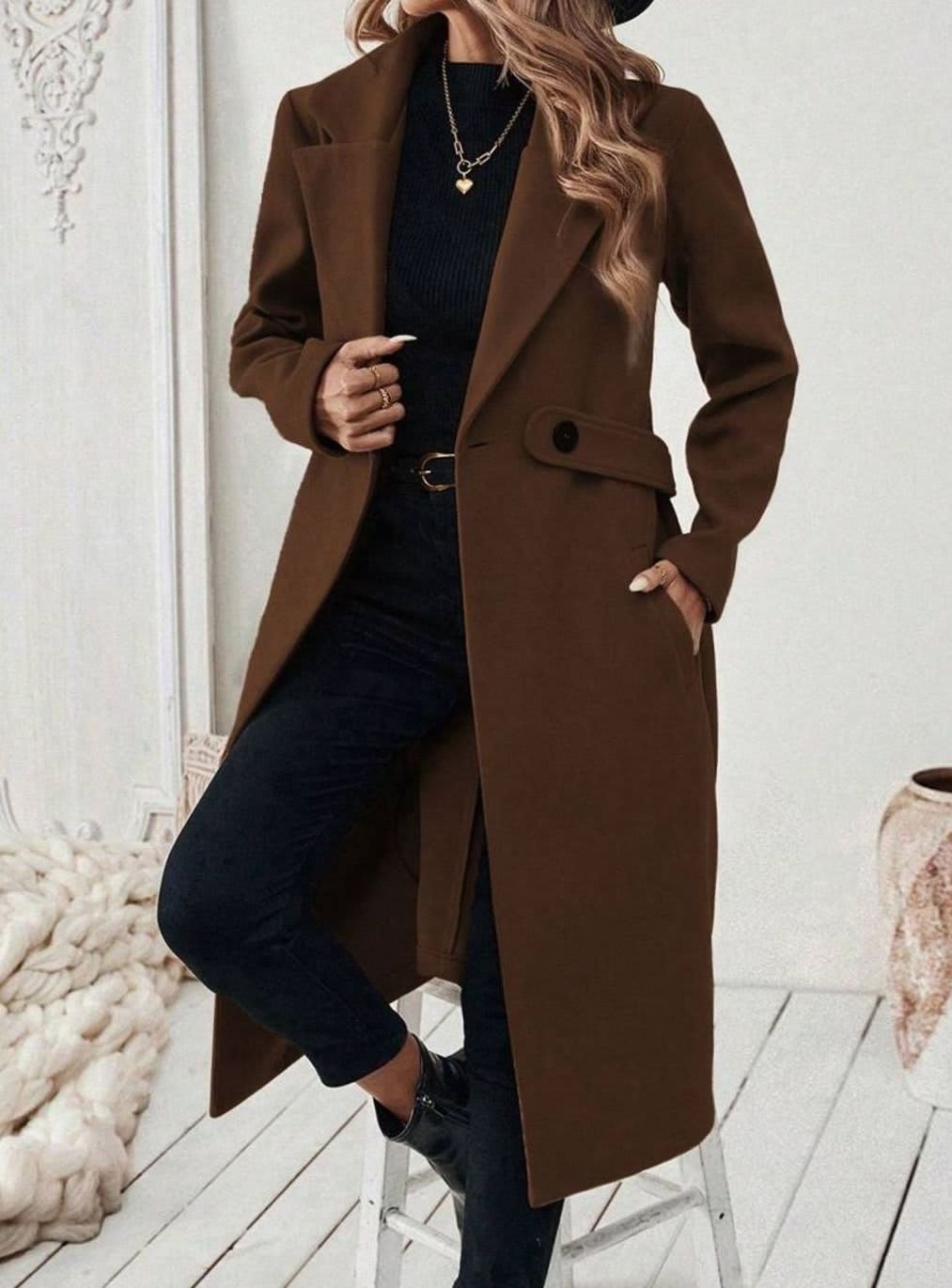 Woman wearing a long notch lapel coat with a buttoned waist tab, welt pockets and long sleeves, holding a quilted handbag._5