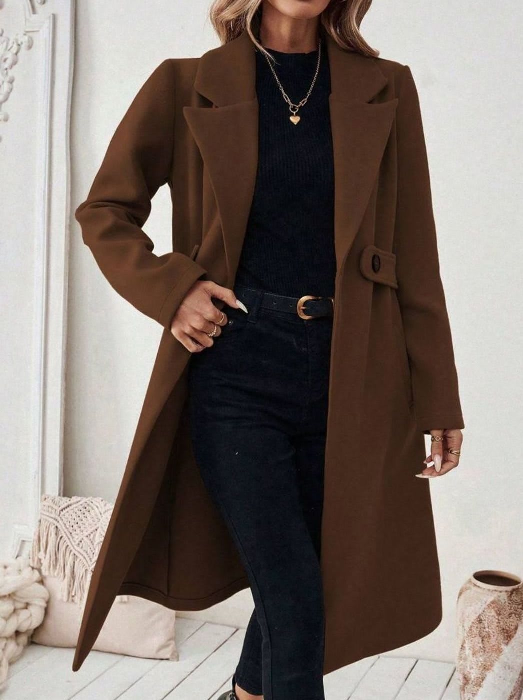 Woman wearing a long notch lapel coat with a buttoned waist tab, welt pockets and long sleeves, holding a quilted handbag._7