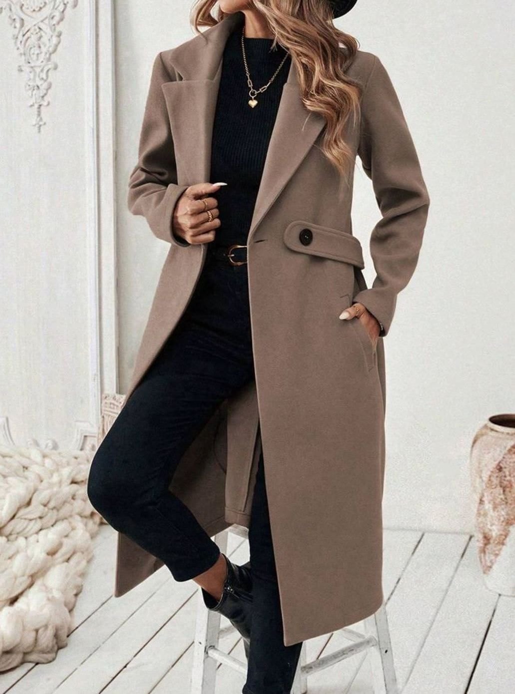Woman wearing a long notch lapel coat with a buttoned waist tab, welt pockets and long sleeves, holding a quilted handbag._9