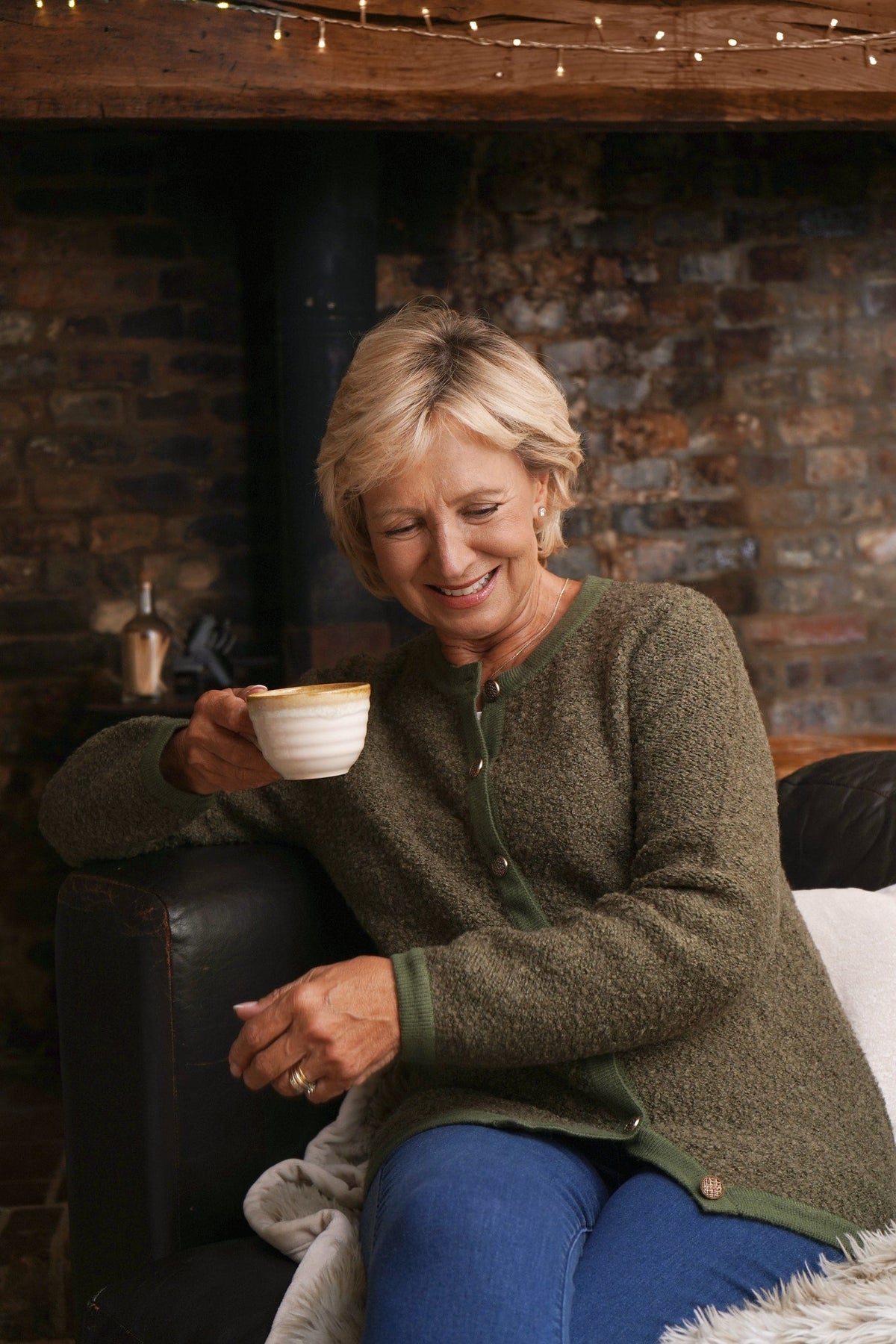 Woman sitting by a brick fireplace holding a cup, wearing a long textured button front cardigan with round neck and ribbed cuffs over jeans._0