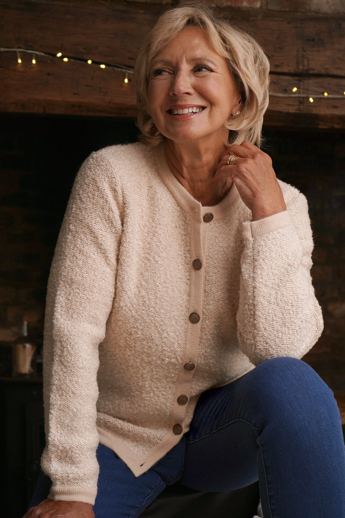 Woman sitting by a brick fireplace holding a cup, wearing a long textured button front cardigan with round neck and ribbed cuffs over jeans._2
