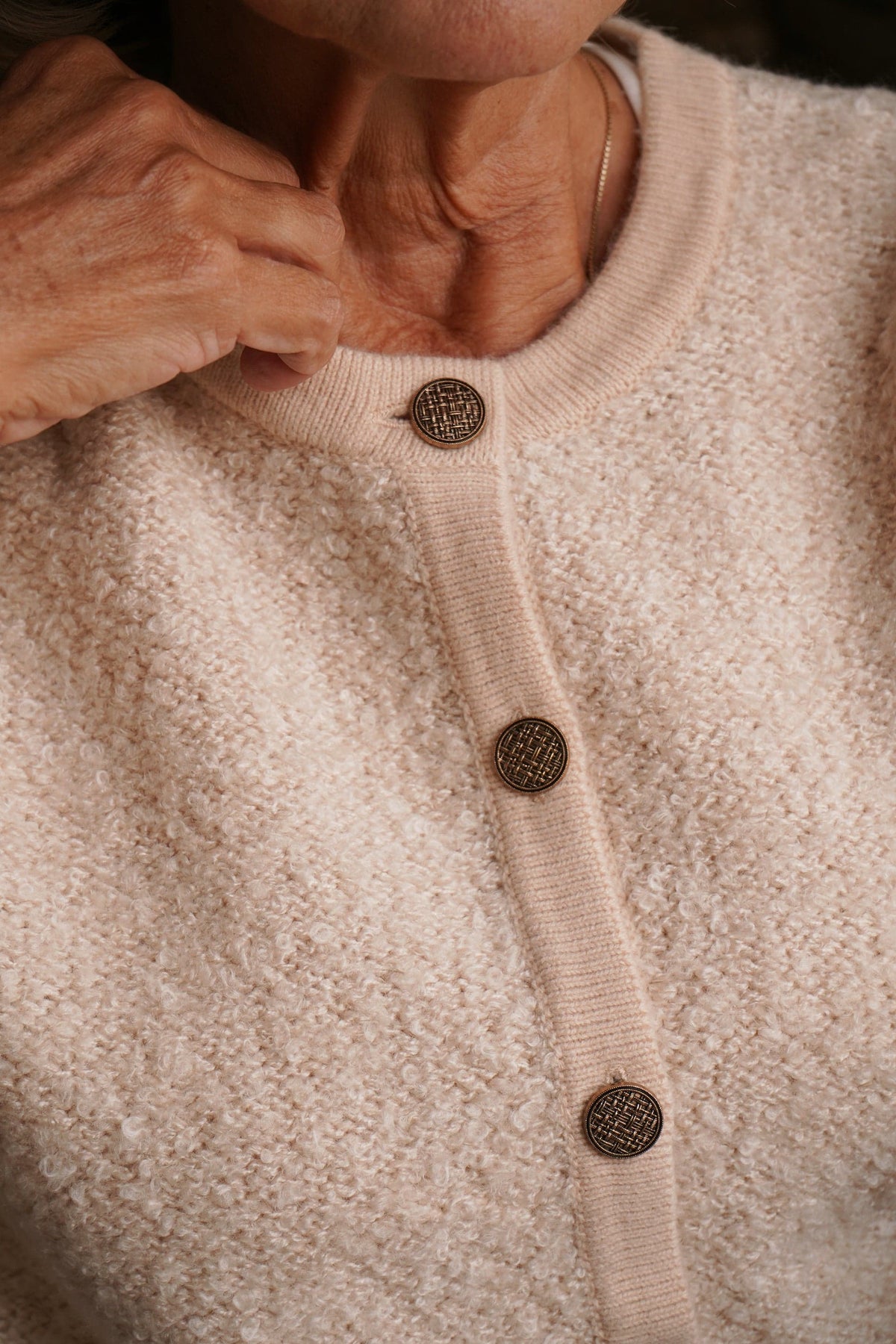 Woman sitting by a brick fireplace holding a cup, wearing a long textured button front cardigan with round neck and ribbed cuffs over jeans._4