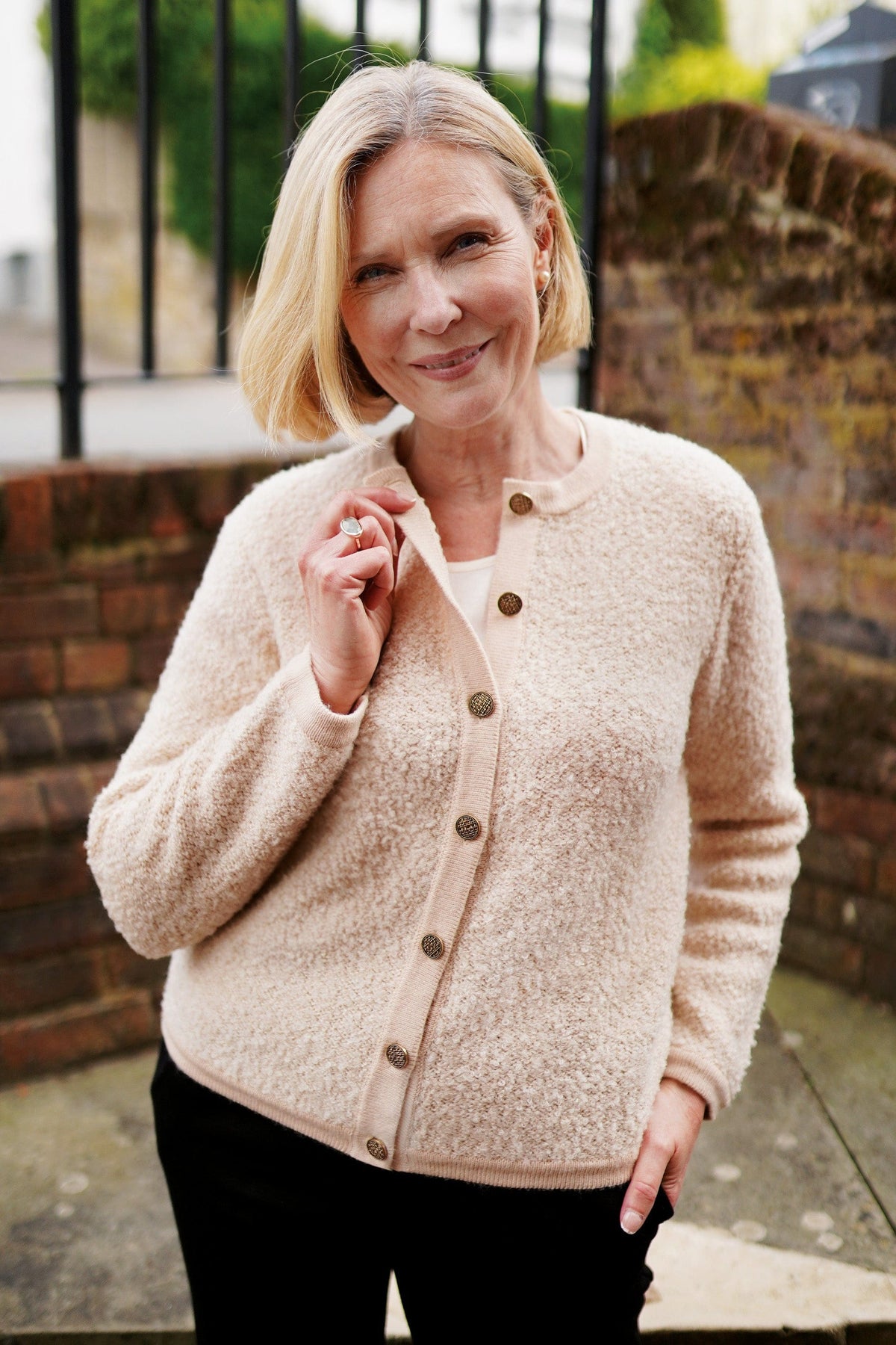 Woman sitting by a brick fireplace holding a cup, wearing a long textured button front cardigan with round neck and ribbed cuffs over jeans._5