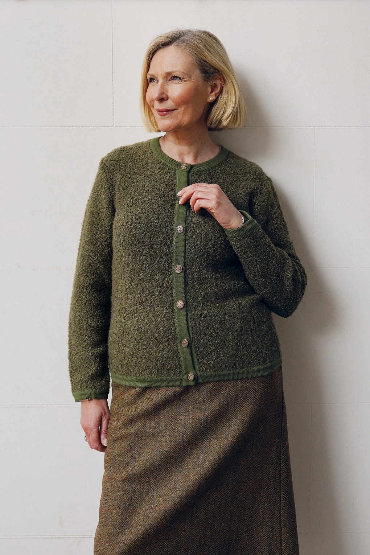 Woman sitting by a brick fireplace holding a cup, wearing a long textured button front cardigan with round neck and ribbed cuffs over jeans._6
