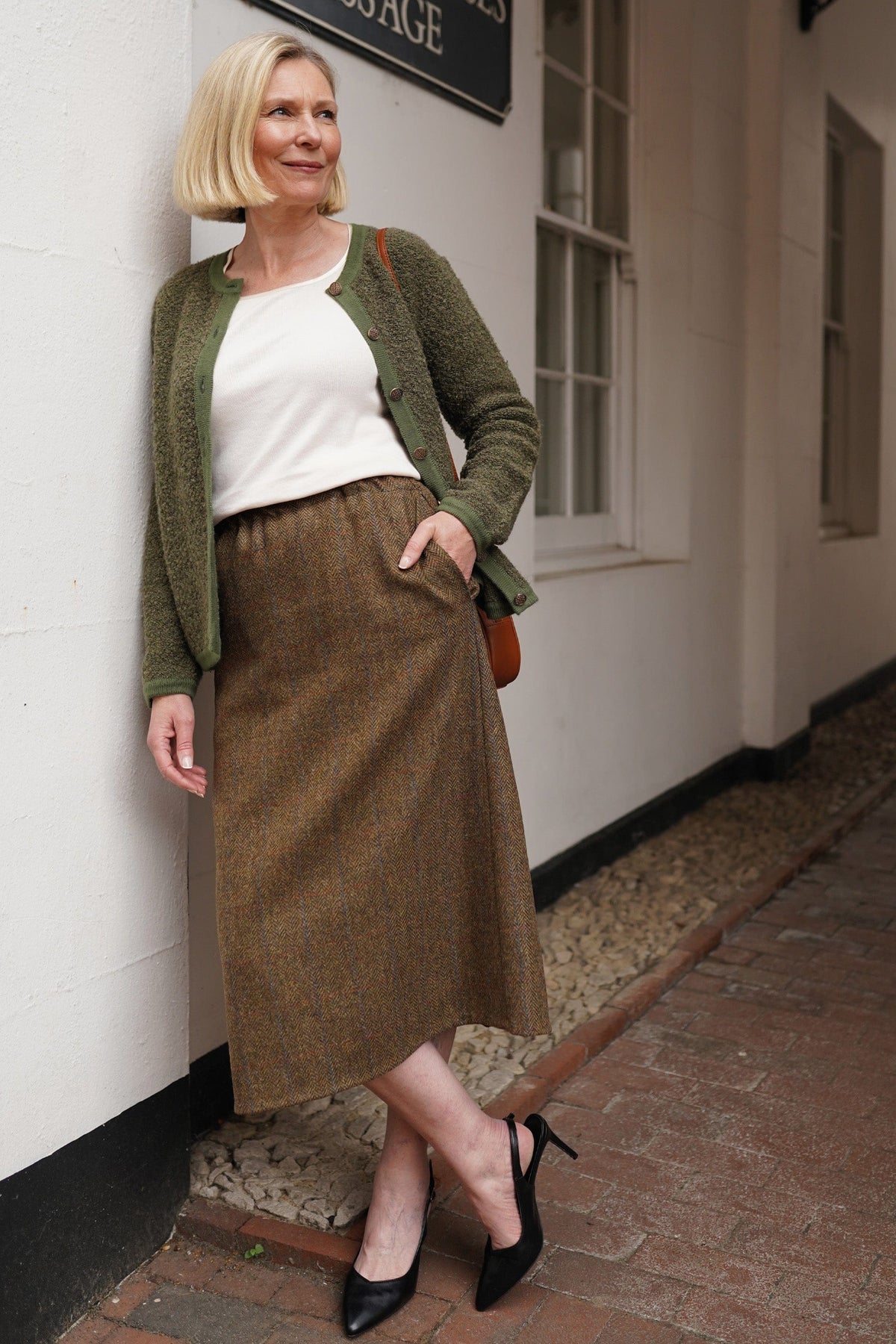 Woman sitting by a brick fireplace holding a cup, wearing a long textured button front cardigan with round neck and ribbed cuffs over jeans._9