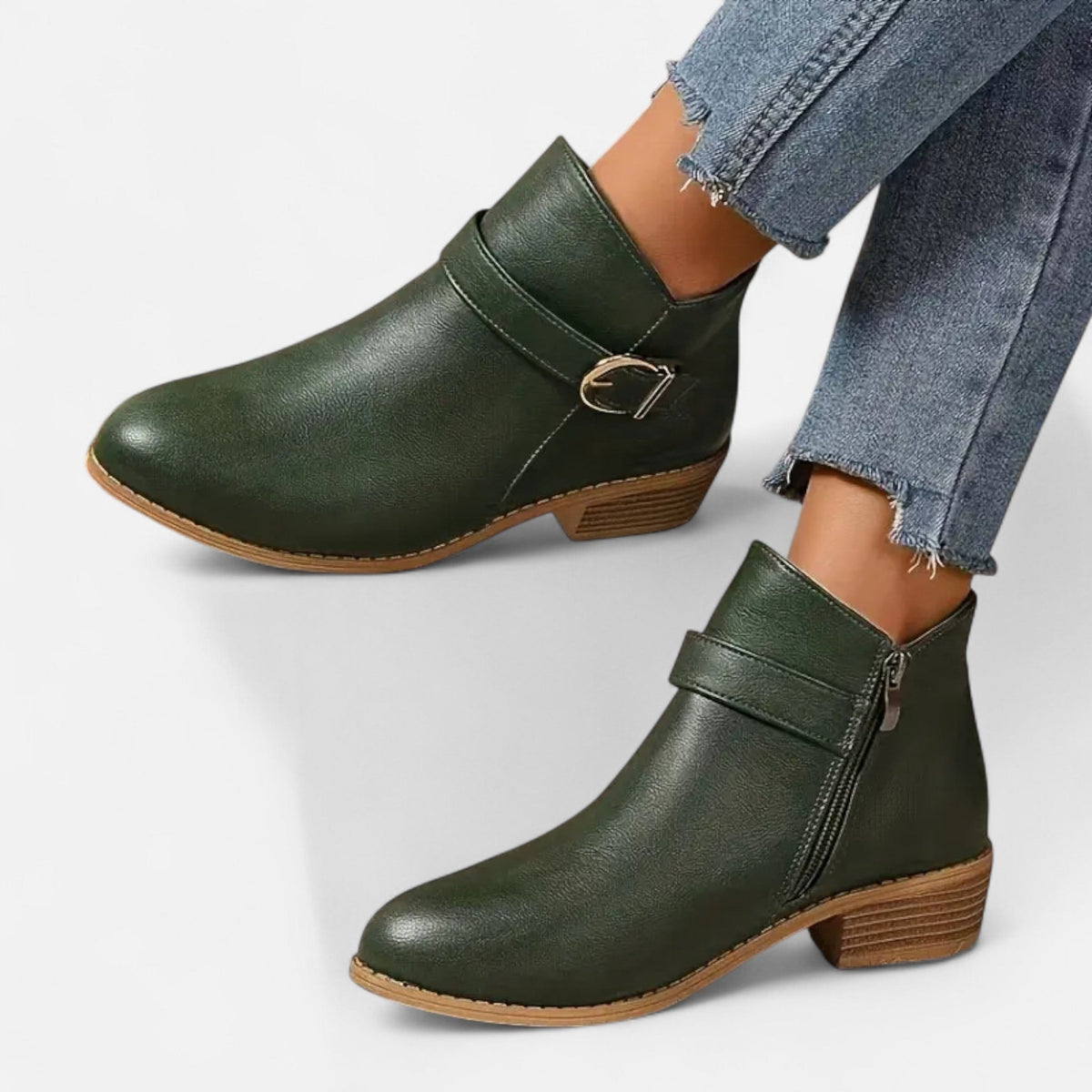 Dark green ankle boots with side buckle straps, visible zippers, tan-edged soles, and low stacked heels worn with jeans.