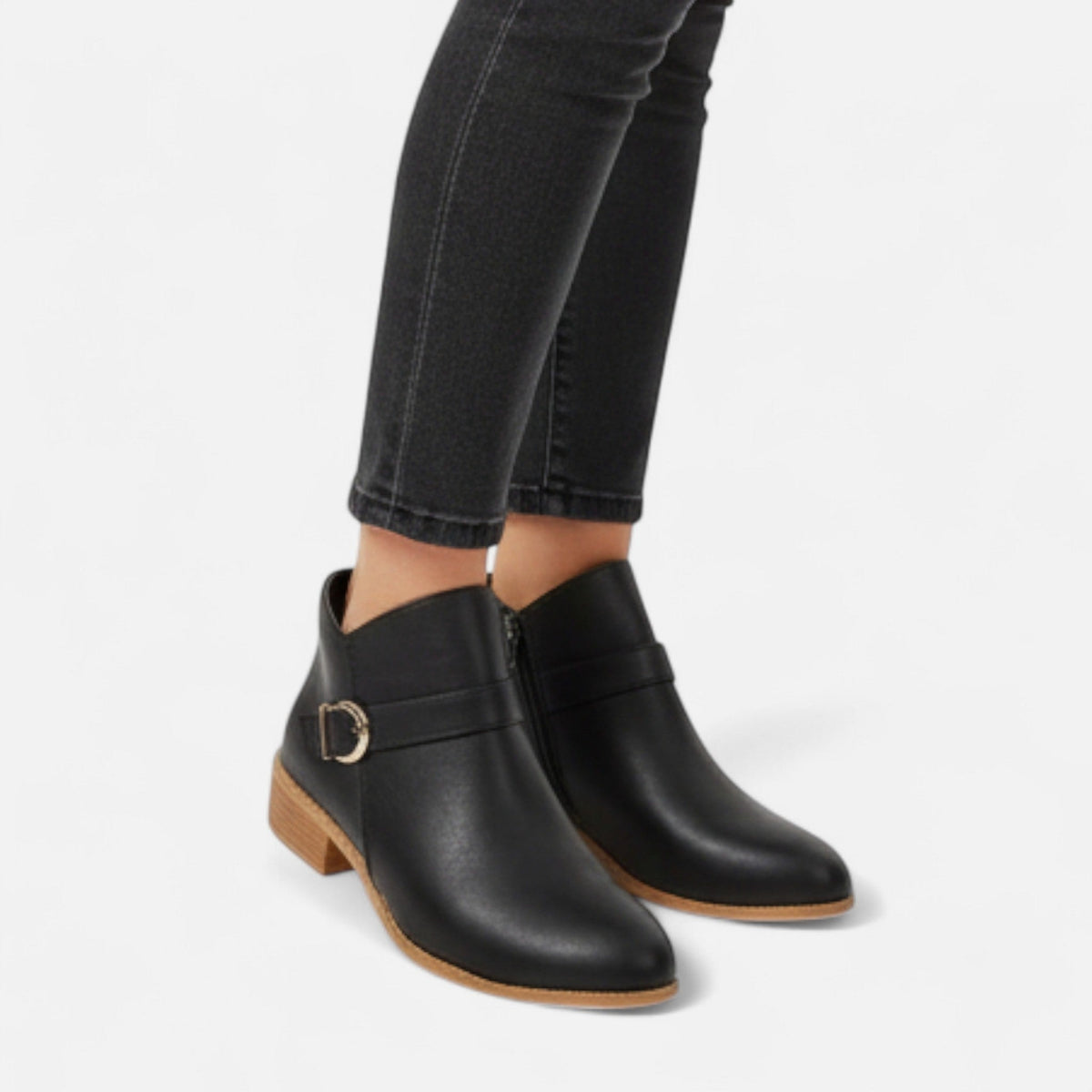 Black ankle boots shown on feet with cropped black jeans, highlighting the side buckle detail and low stacked heels.