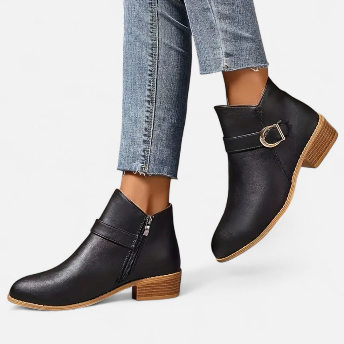 Black buckle strap ankle boots shown from the side with one boot lifted, revealing the zipper, textured stacked heel, and tan-edged sole.