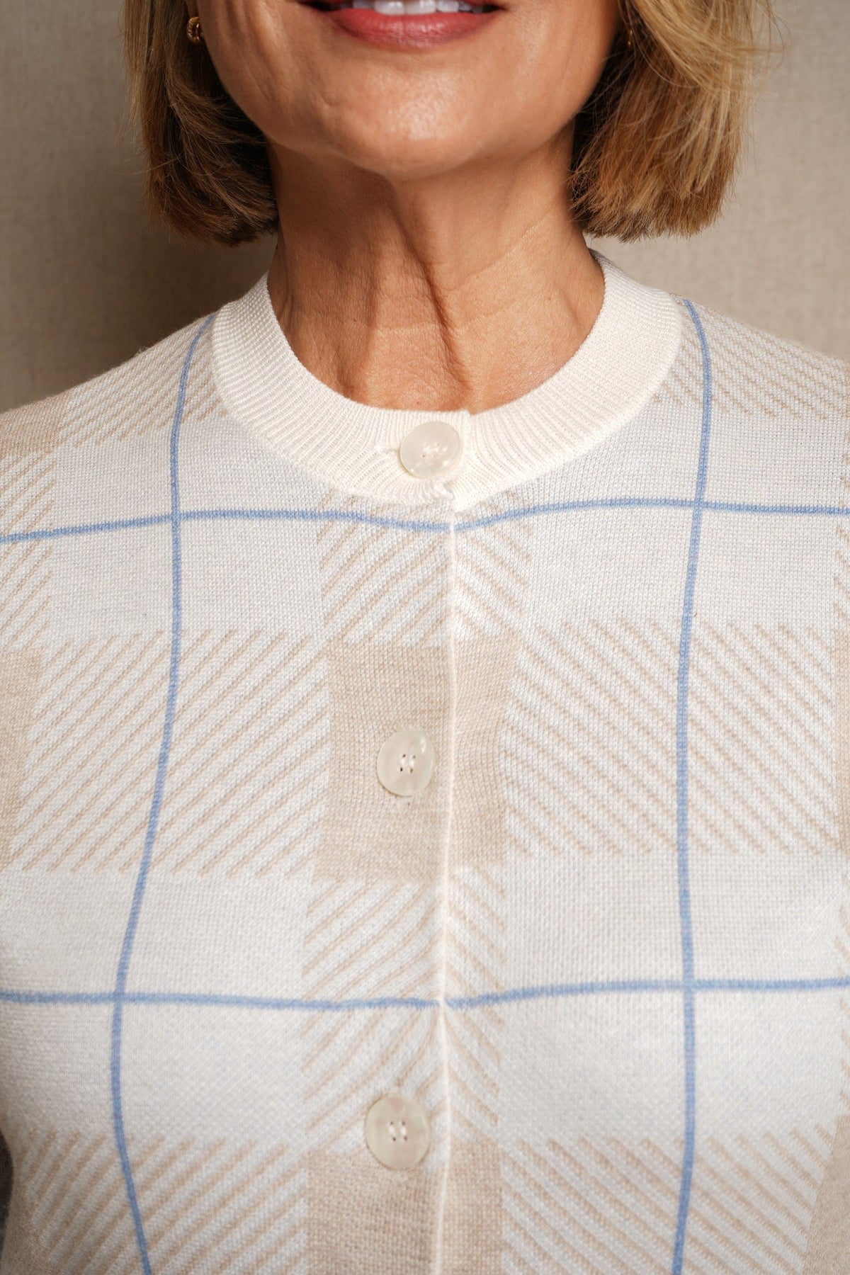 Woman seated wearing a long sleeve button front knit cardigan with a large check pattern and ribbed crew neck, cuffs and hem, with jeans._2