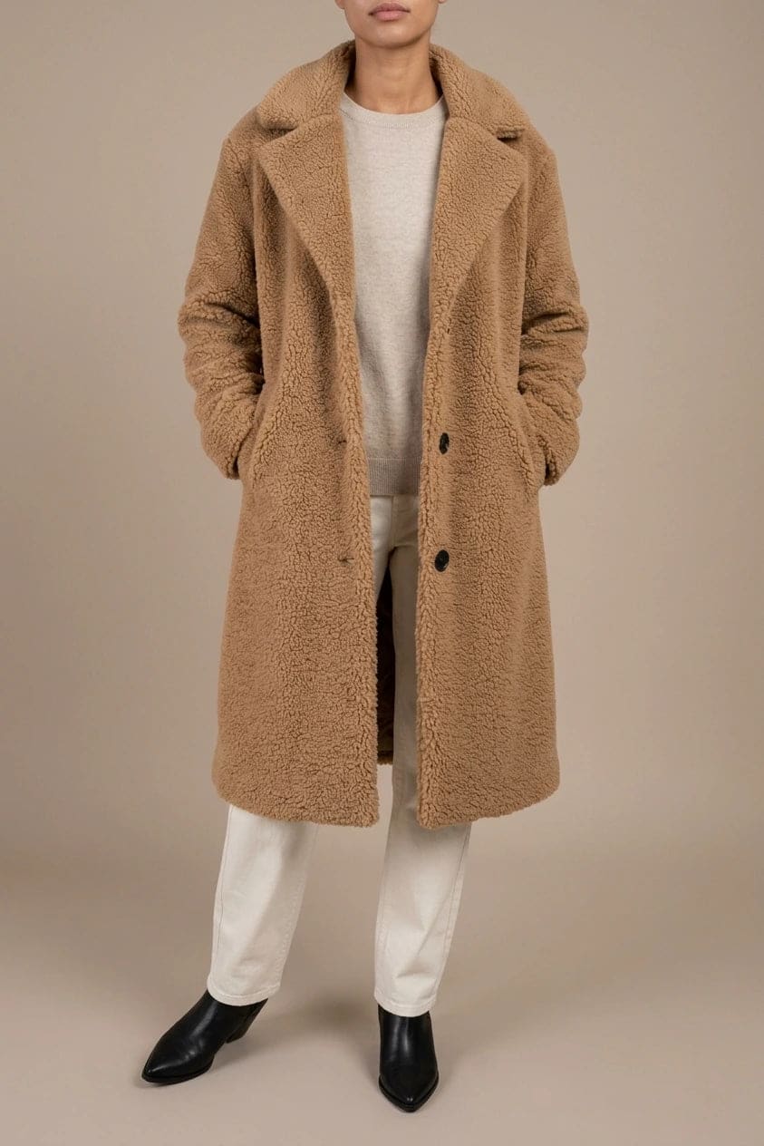 Woman wearing a long textured teddy coat with notch lapels and button front, open over a jumper and trousers with ankle boots._0