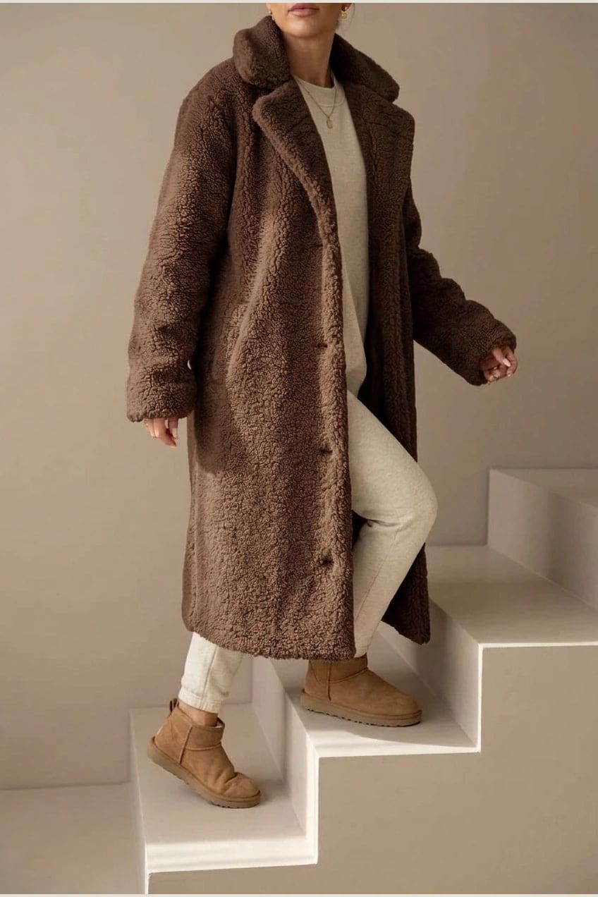 Woman wearing a long textured teddy coat with notch lapels and button front, open over a jumper and trousers with ankle boots._11