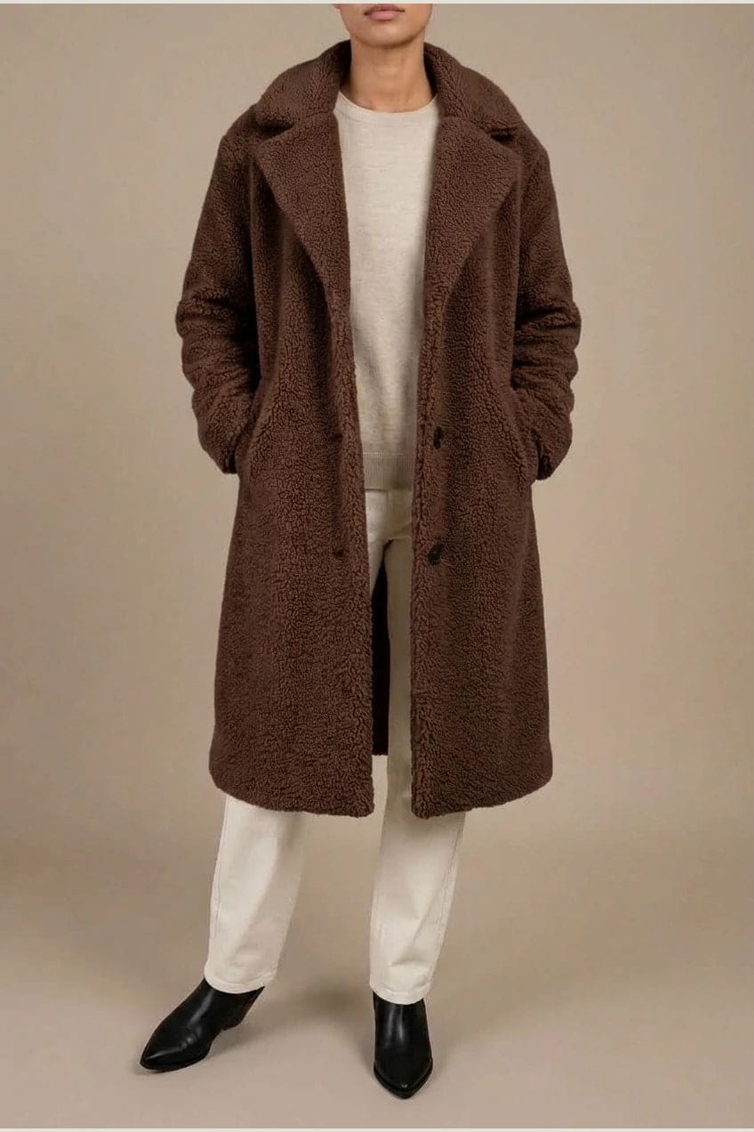 Woman wearing a long textured teddy coat with notch lapels and button front, open over a jumper and trousers with ankle boots._7