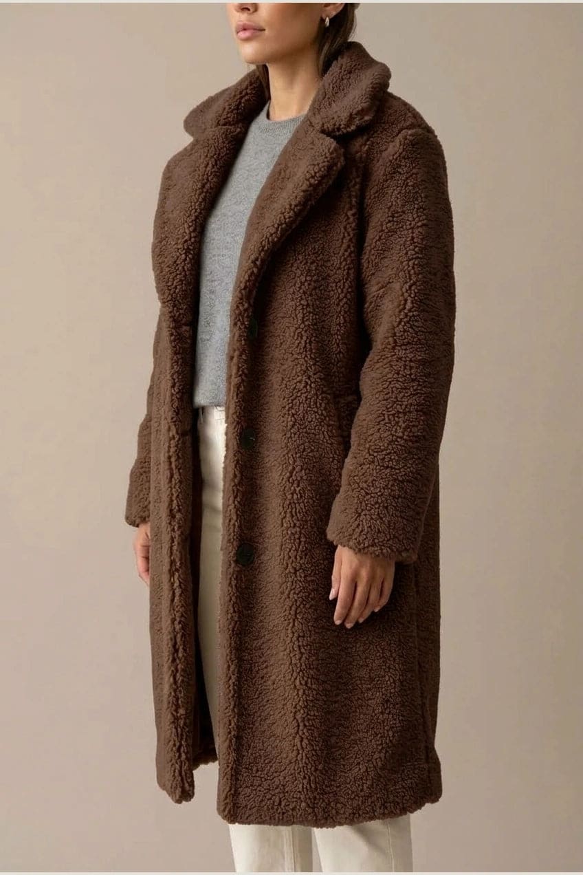 Woman wearing a long textured teddy coat with notch lapels and button front, open over a jumper and trousers with ankle boots._8