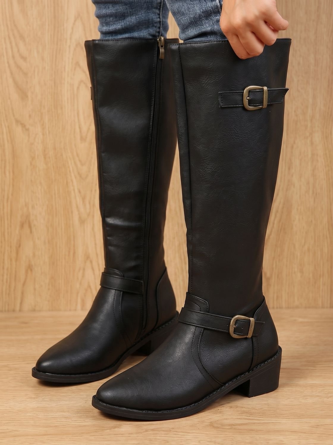 Black knee-high boots with two buckle accents, side zips, pointed toes, and low block heels shown with jeans.