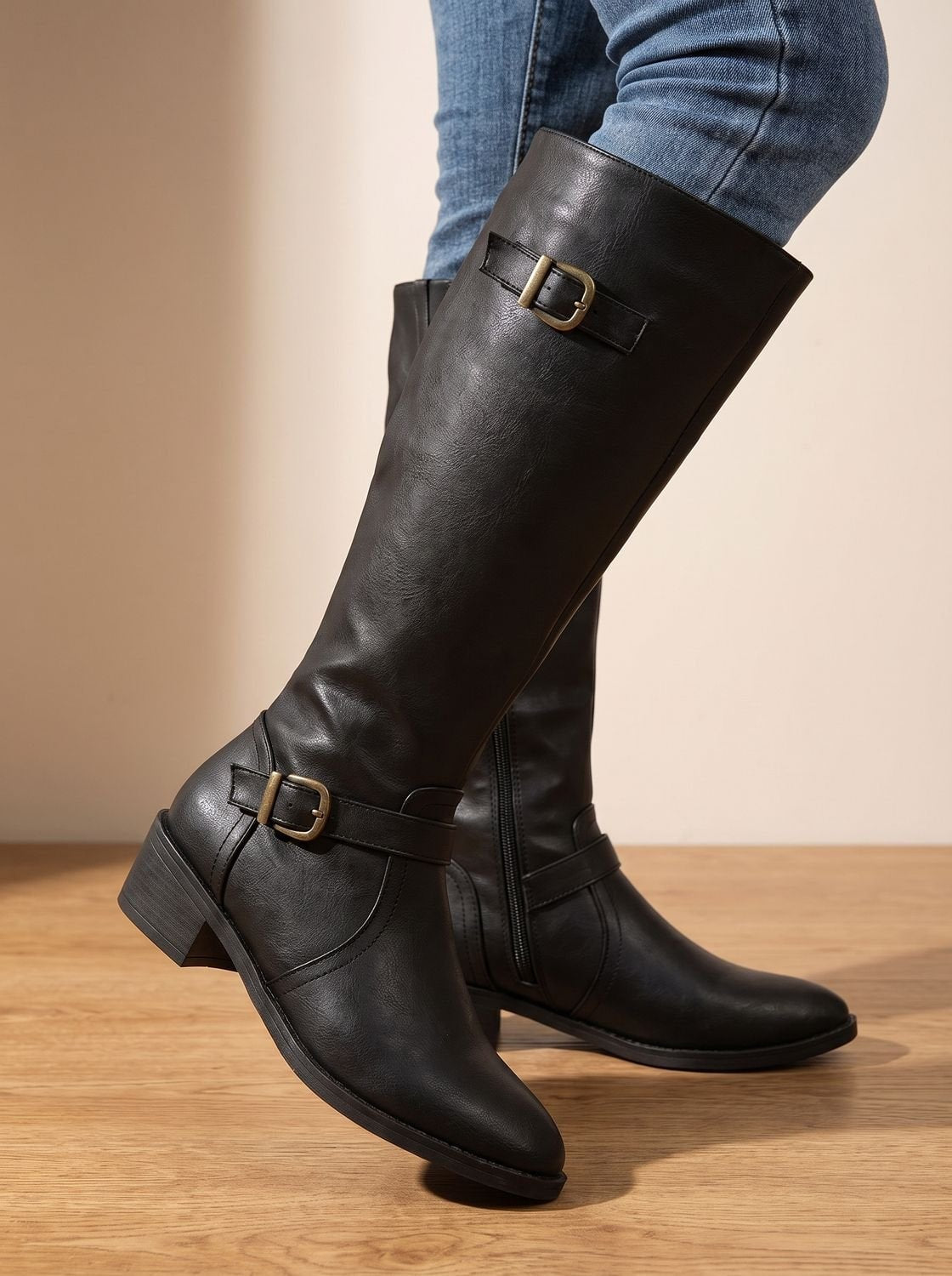 Black knee-high boots shown crossed at the ankles, highlighting the side profile, upper buckle, ankle buckle, and block heel.