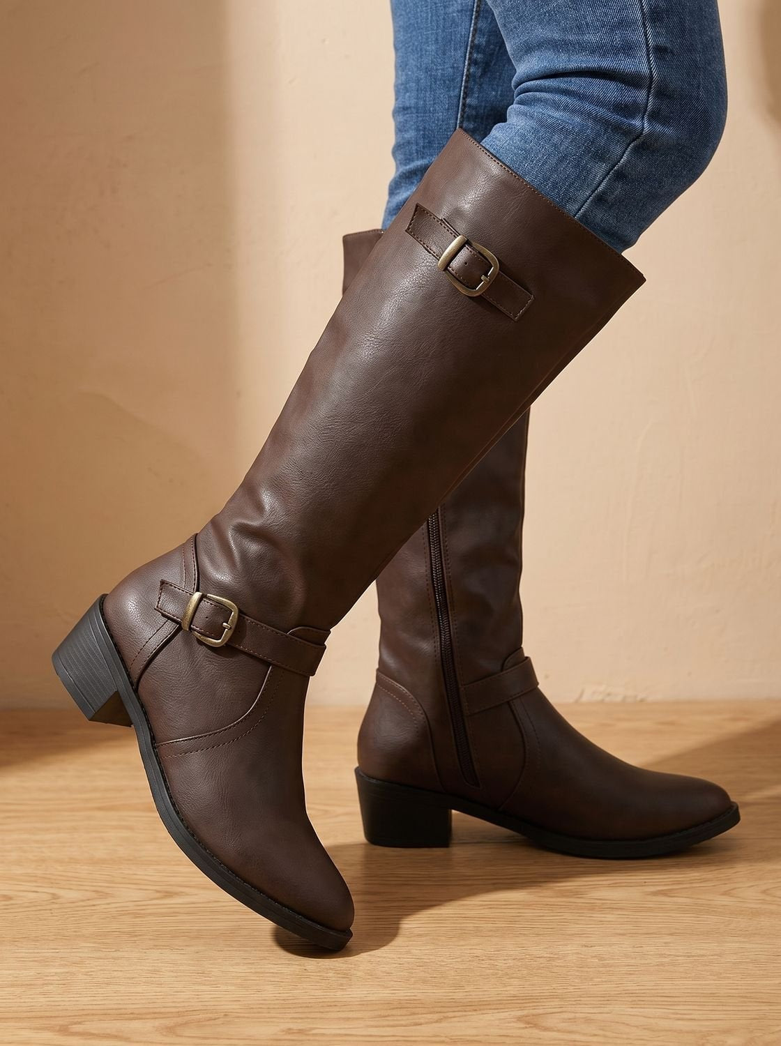 Brown knee-high boots displayed in a crossed-leg pose, showing the rounded pointed toes, two buckles, side zip, and low heel.