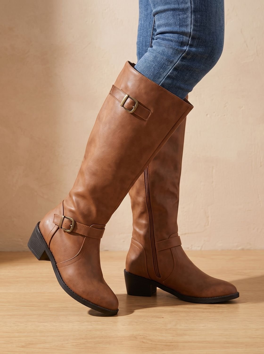 Tan brown knee-high boots shown in side view with contrasting buckle accents, visible side zips, and sturdy low heels.