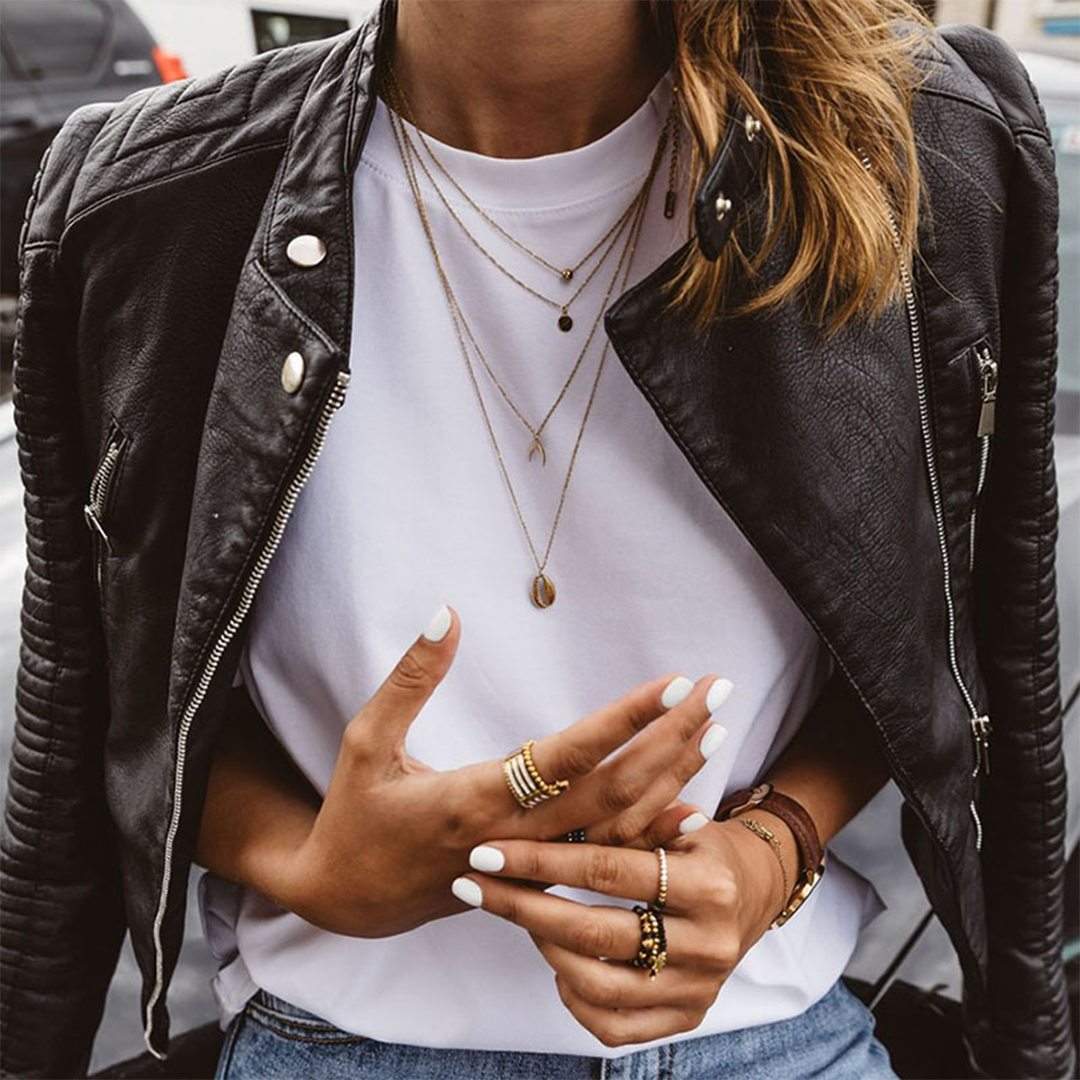 Close view of the black biker jacket layered over a white shirt, highlighting the open asymmetric front, snap-tab collar, metallic zips, and quilted shoulders and sleeves.
