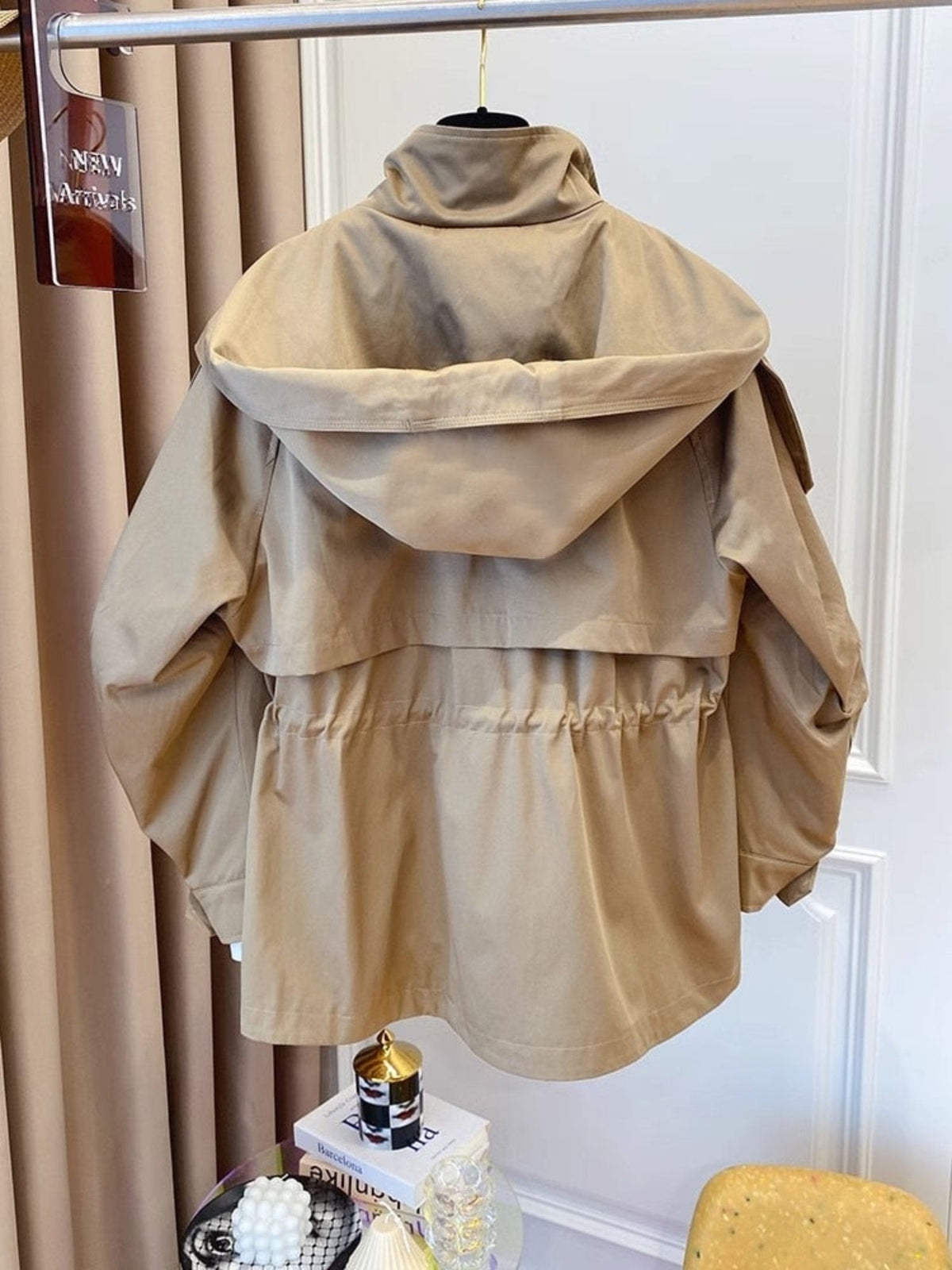 Back view of the beige jacket on display, highlighting the broad hood, layered back panel, cinched drawstring waist, and curved hem.