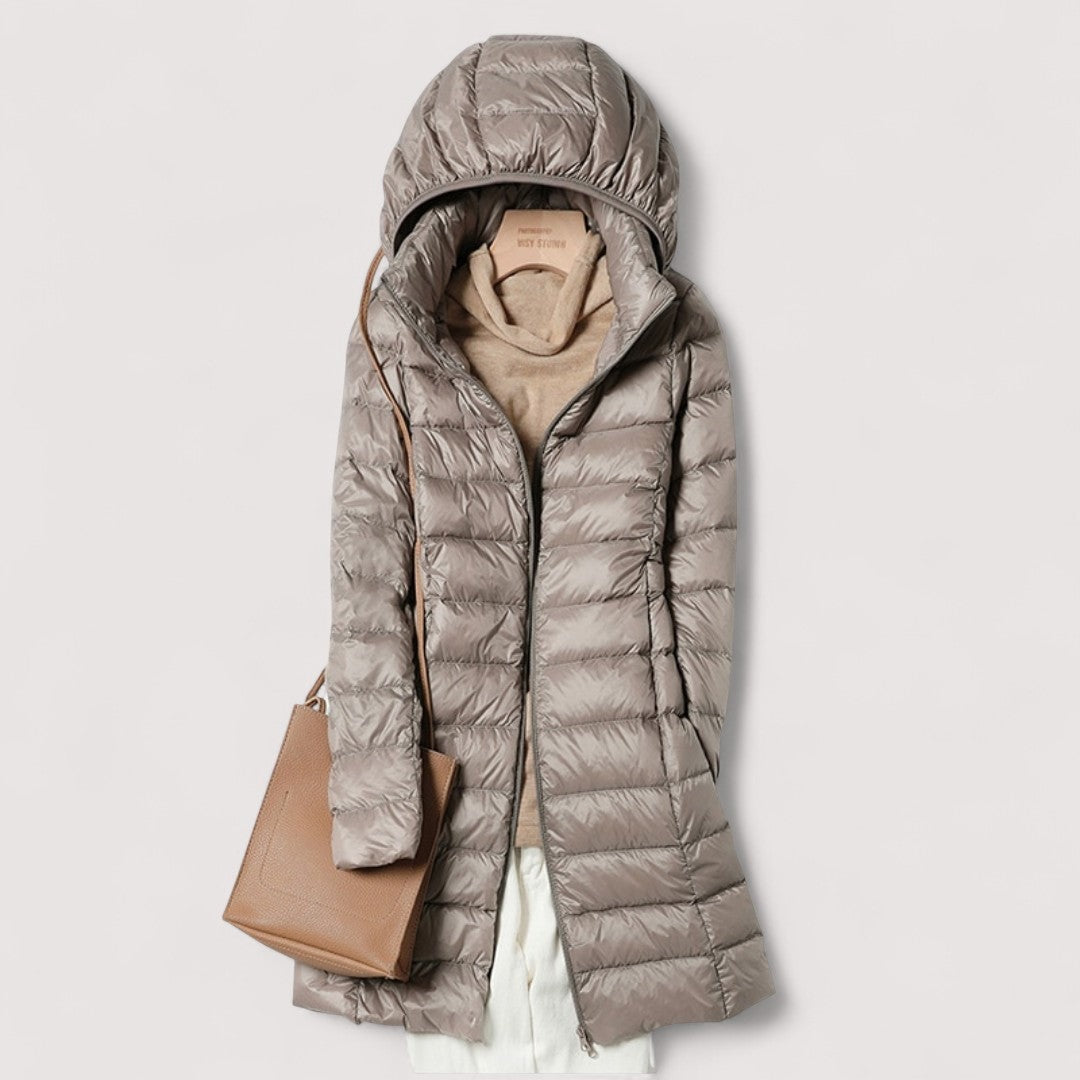 Taupe hooded longline puffer jacket shown zipped over a beige top, paired with a tan tote bag.