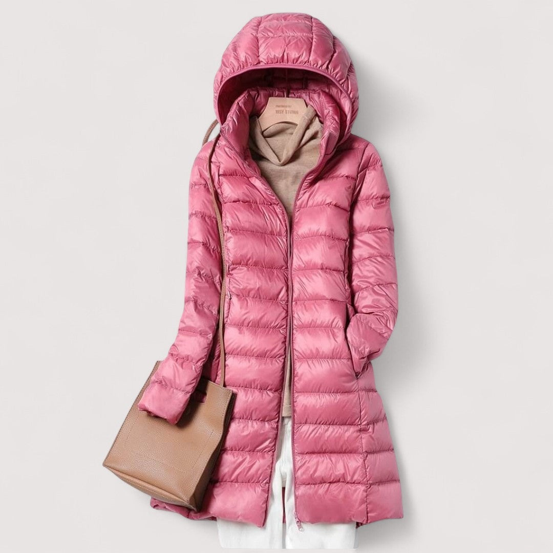 Bright pink hooded longline puffer jacket styled over light clothing, with a tan shoulder bag.